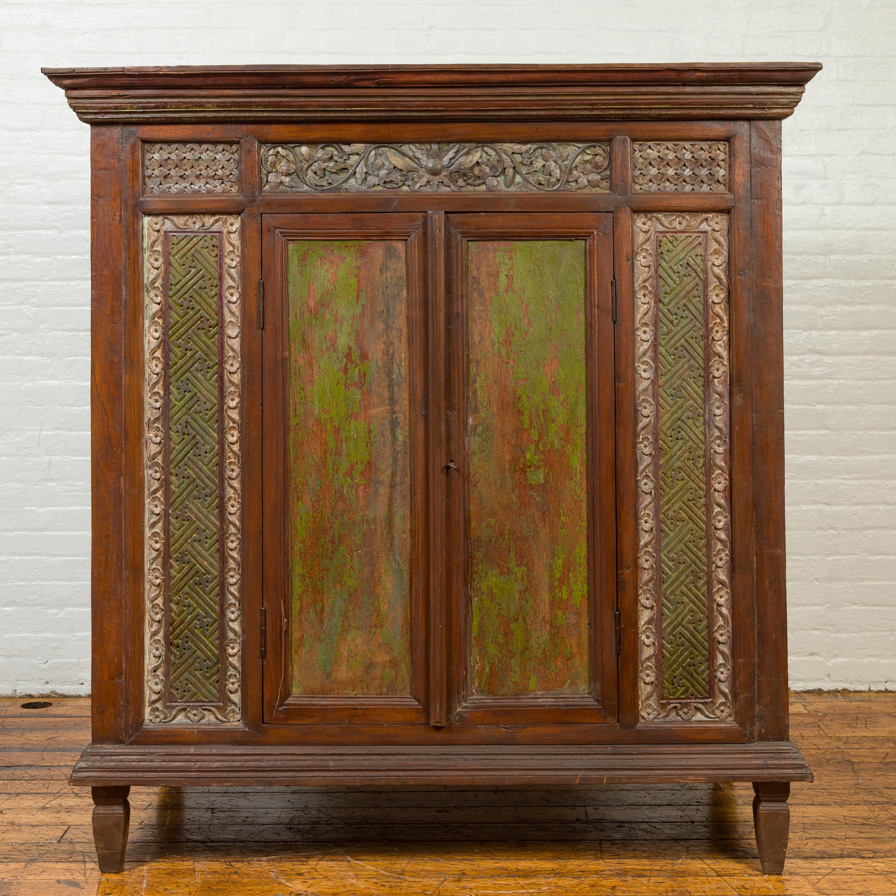 A large antique Indonesian cabinet from the 19th century, with carved floral decor and hand painted distressed decor. Crafted in Indonesia during the 19th century, this large cabinet features a molded cornice overhanging a richly carved façade