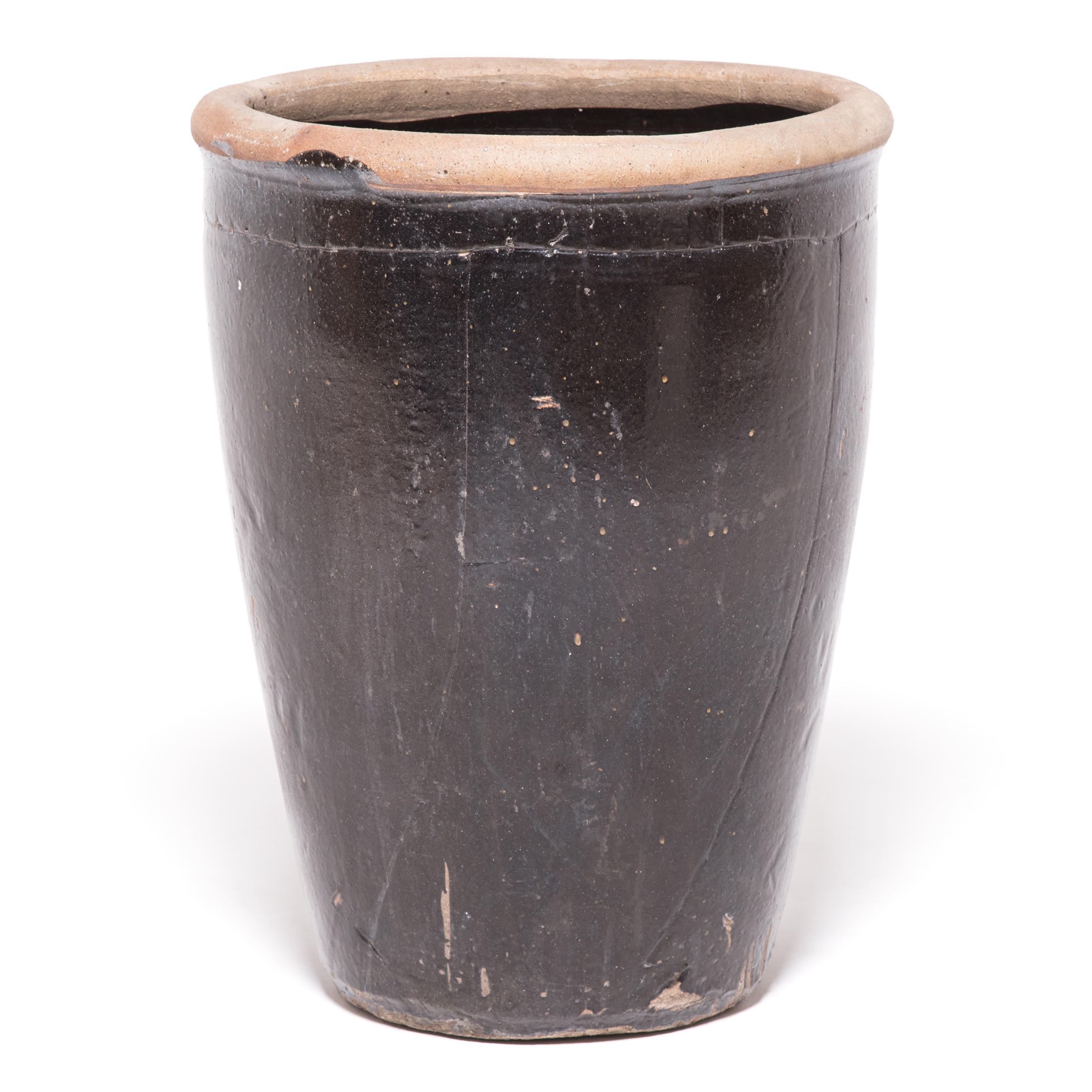 Originally used for pickling and storing various foods, this tall ceramic jar features a strikingly dark glaze. Set in high contrast by an unglazed lip and speckled with imperfections, the glazed surface exudes a rich luster and makes this jar the