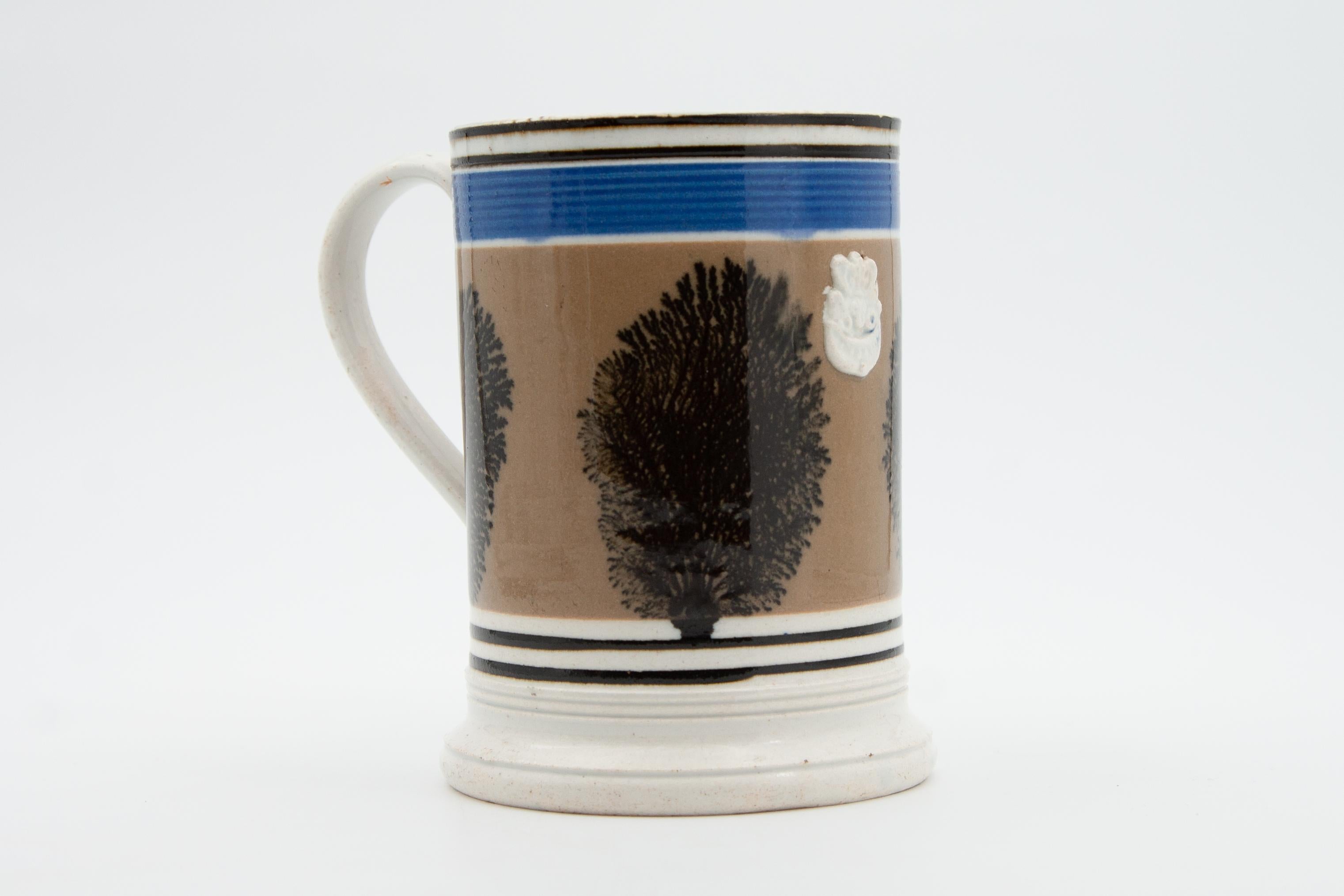 An English Mochaware mug with dendritic or tree decoration, dating to circa 1840.

This large Mochaware mug features rouletted bands of black and blue framing a tree-like pattern (referred to as “algae” or “dendrite”) on a taupe brown background.