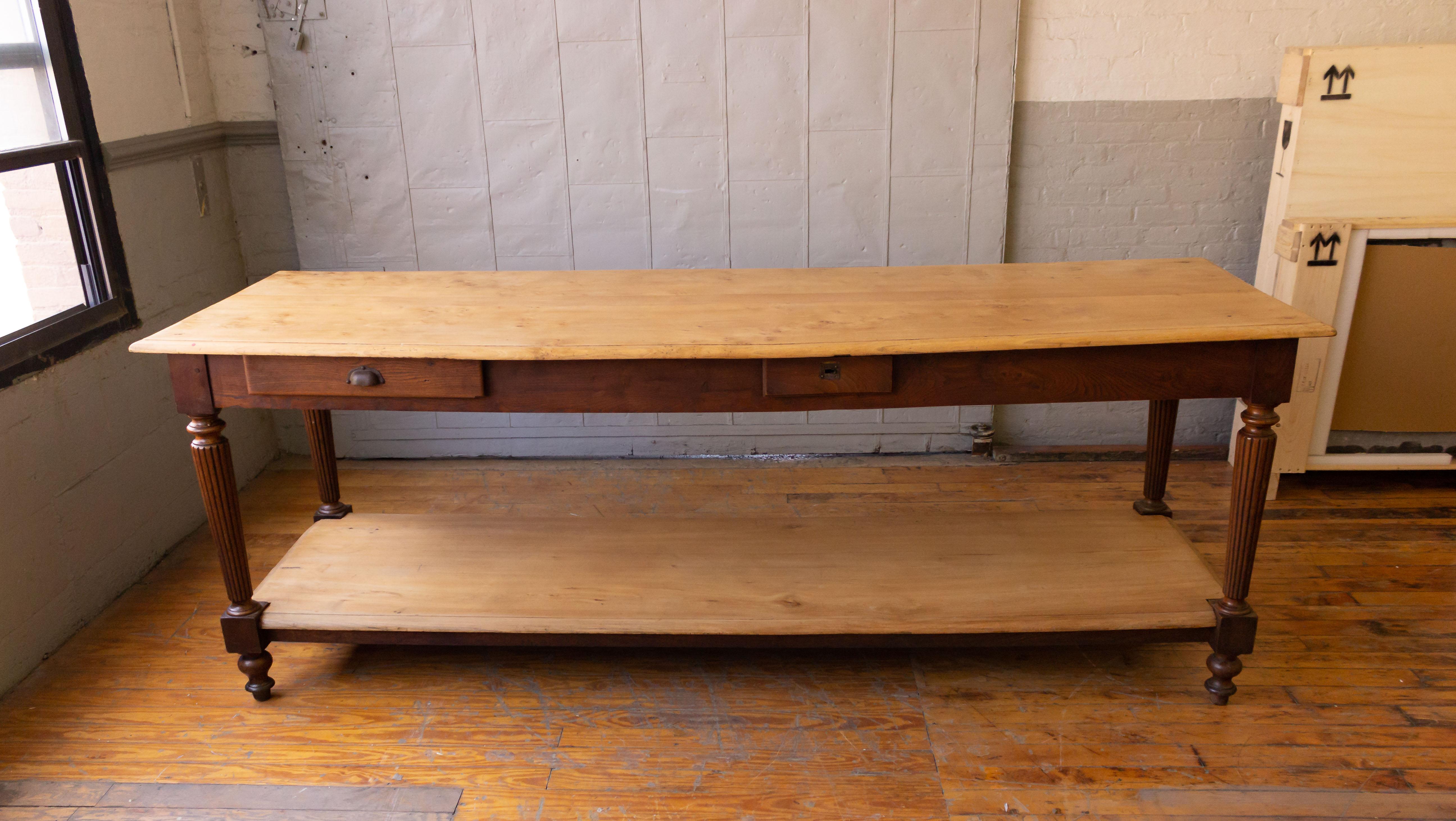 An interesting French late 19th century or early 20th century draper's table made in Elm. The upper and lower shelves are in natural wood finish and the legs are stained in darker wood tones. The top has a slight bow, but the table is structurally