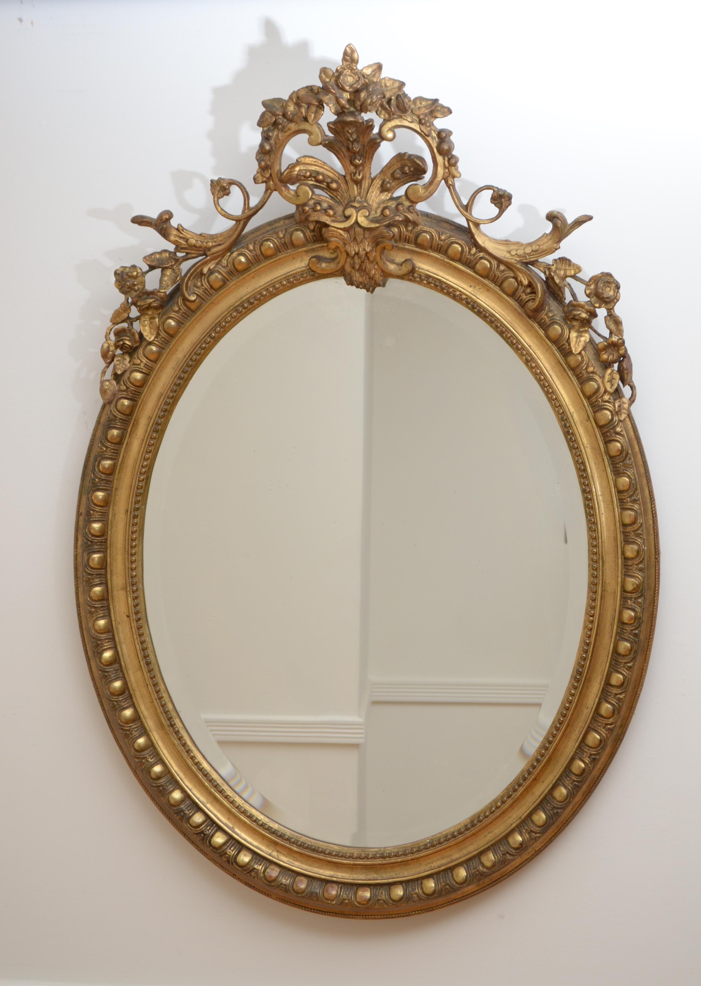 K0066 a superb gilded mirror of oval from, having original bevelled edge glass with some foxing in beautifully beaded and egg and dart carved giltwood frame with floral centre crest to the top. This antique wall mirror retains its original glass,
