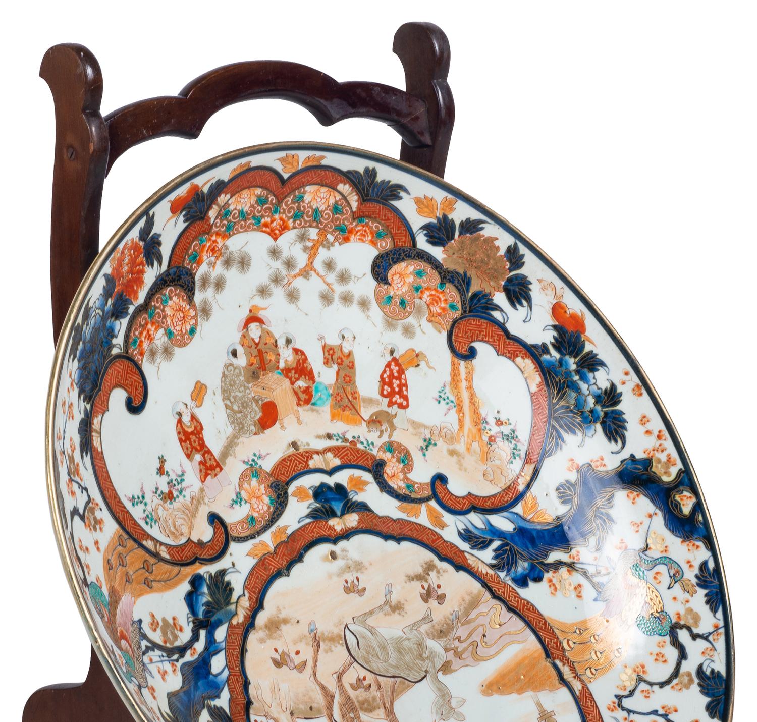 An impressive good quality 19th century Japanese Imari charger on a 19th century mahogany plate stand.
The charger having classical exotic bird and foliate decoration, inset hand painted panels depicting courtiers playing games and deer under trees.