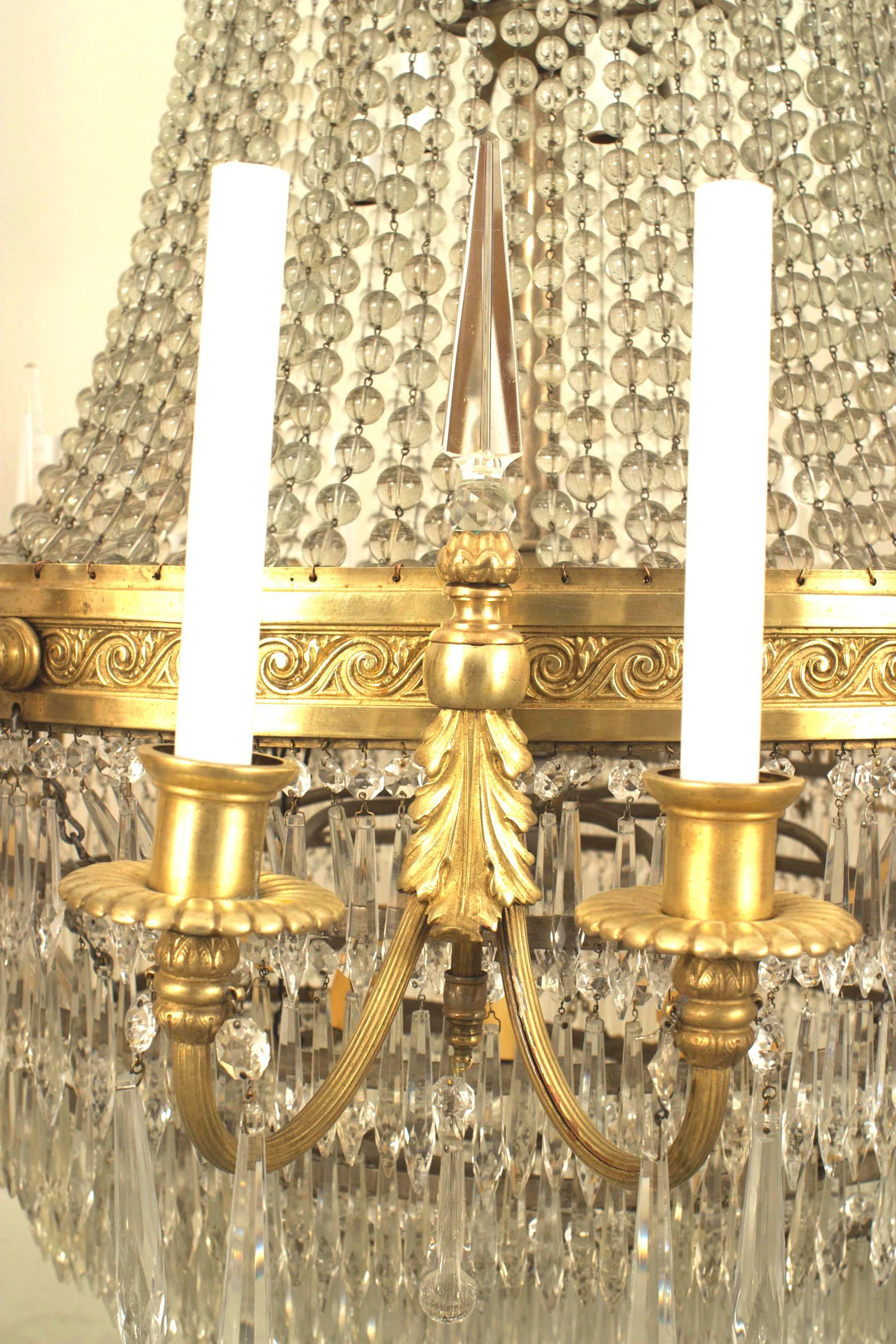 Grand lustre en cristal de style Louis XVI (20e siècle) avec un anneau en bronze doré supportant 24 bras à volutes avec des lumières sous des rangées de cristaux perlés avec des prismes à gouttes en cristal à plusieurs niveaux en dessous.
