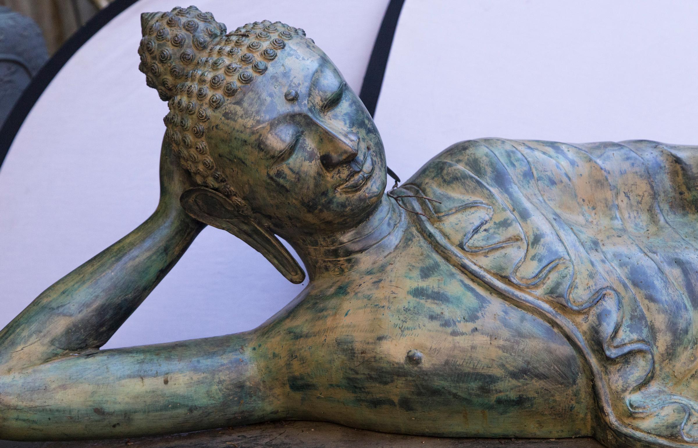 The bronze figure reclines in typical pose, with his head propped up on his right arm and hand. He wears a loose garment. Elongated ear lobes. Can be used outdoors or indoors.