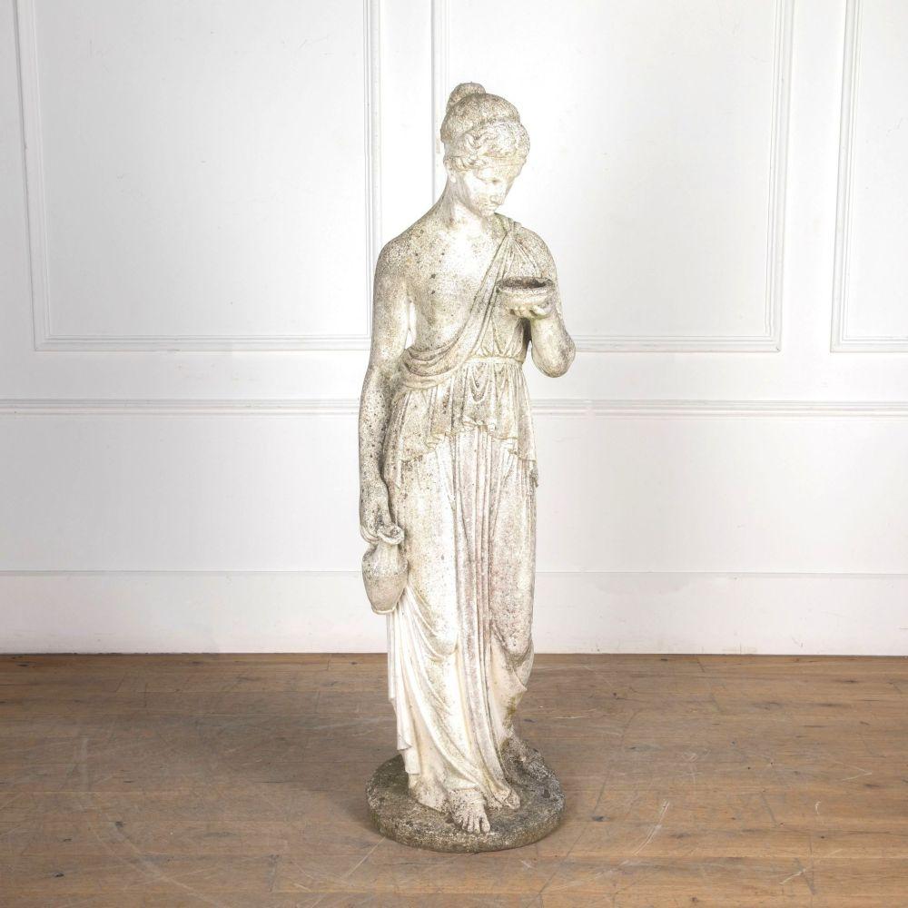A weathered reconstituted marble statue of Hebe in classical robes holding a cup. 

Hebe is a Goddess in Greek mythology, a daughter of Zeus and cupbearer to the Gods. She is also associated with the quest for eternal youth. 

This figure is made
