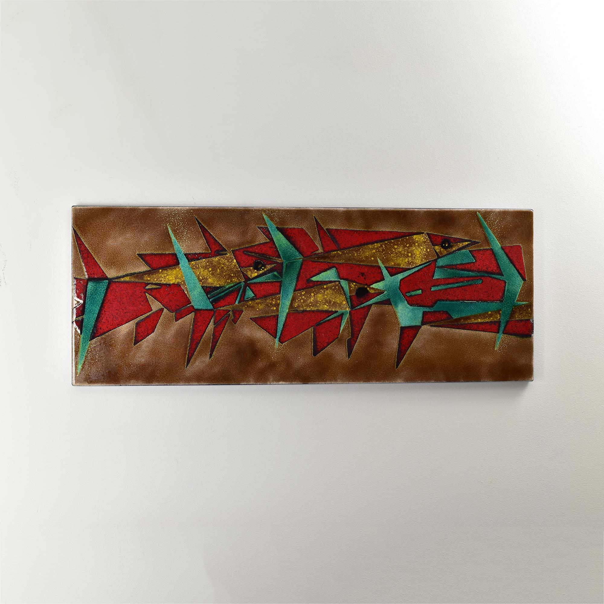 Cette plaque murale émaillée du milieu du siècle présente un motif géométrique audacieux de poisson, rappelant l'influence artistique de Gio Ponti. Présentant des teintes vibrantes de rouge, de turquoise et d'or sur un fond marron texturé, le design