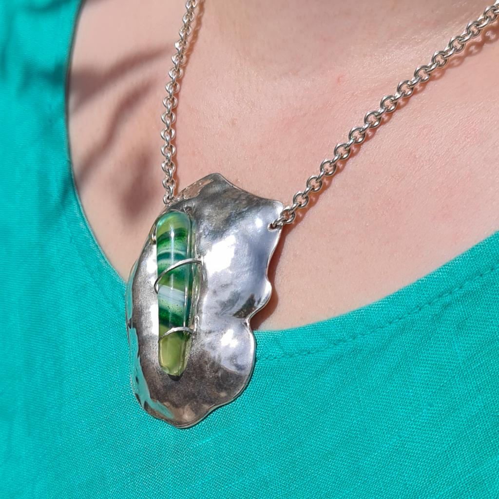 This statement large contemporary abstract silver pendant has been chased by hand using traditional repoussé techniques into an organic fluid form representing the contours of a hill in Gozo, Malta. A fragment of vibrant green Mdina Glass is set
