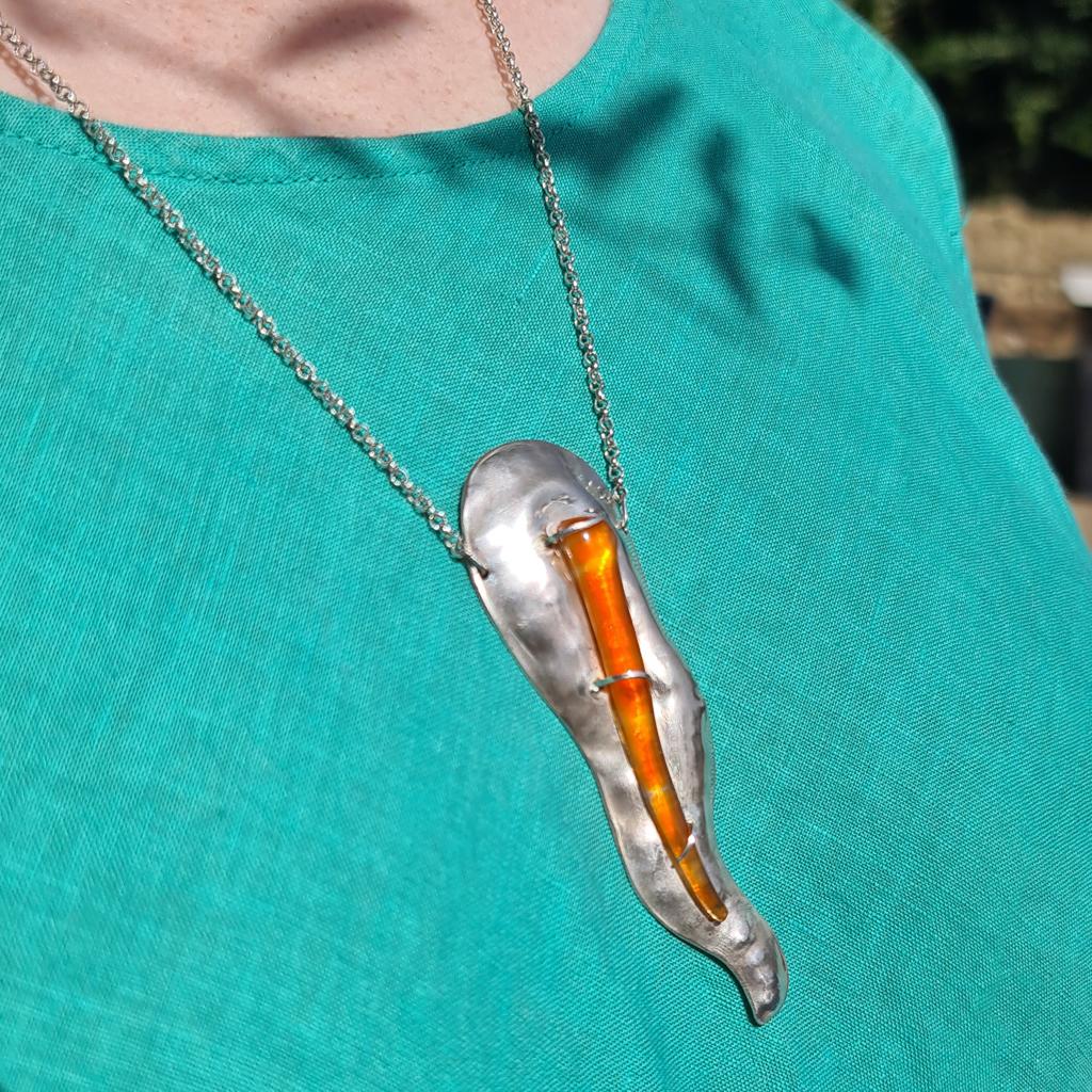 This large contemporary abstract silver pendant has been chased by hand into an organic fluid form representing the contours of a hill in Gozo, Malta. A fragment of ombré orange Mdina Glass, with streaks of yellow, is set like a