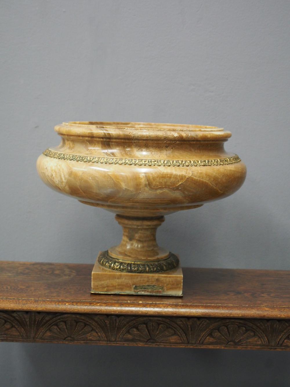 Roman style large Alabaster footed urn. The top and central aperture is unpolished and rises up to a raised for edge. There is a gilded section near the top and it is on a stem with foliate casting before finishing on a square block with brass