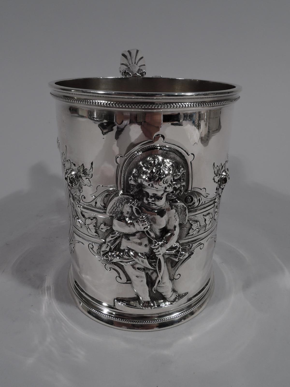 Large American classical coin silver baby cup. Made by Wood & Hughes in New York, ca 1860. Upward tapering sides with skirted foot and scroll-bracket handle. Jowly, tousled-haired contrapposto cherub applied to front. Two oval frames and one small