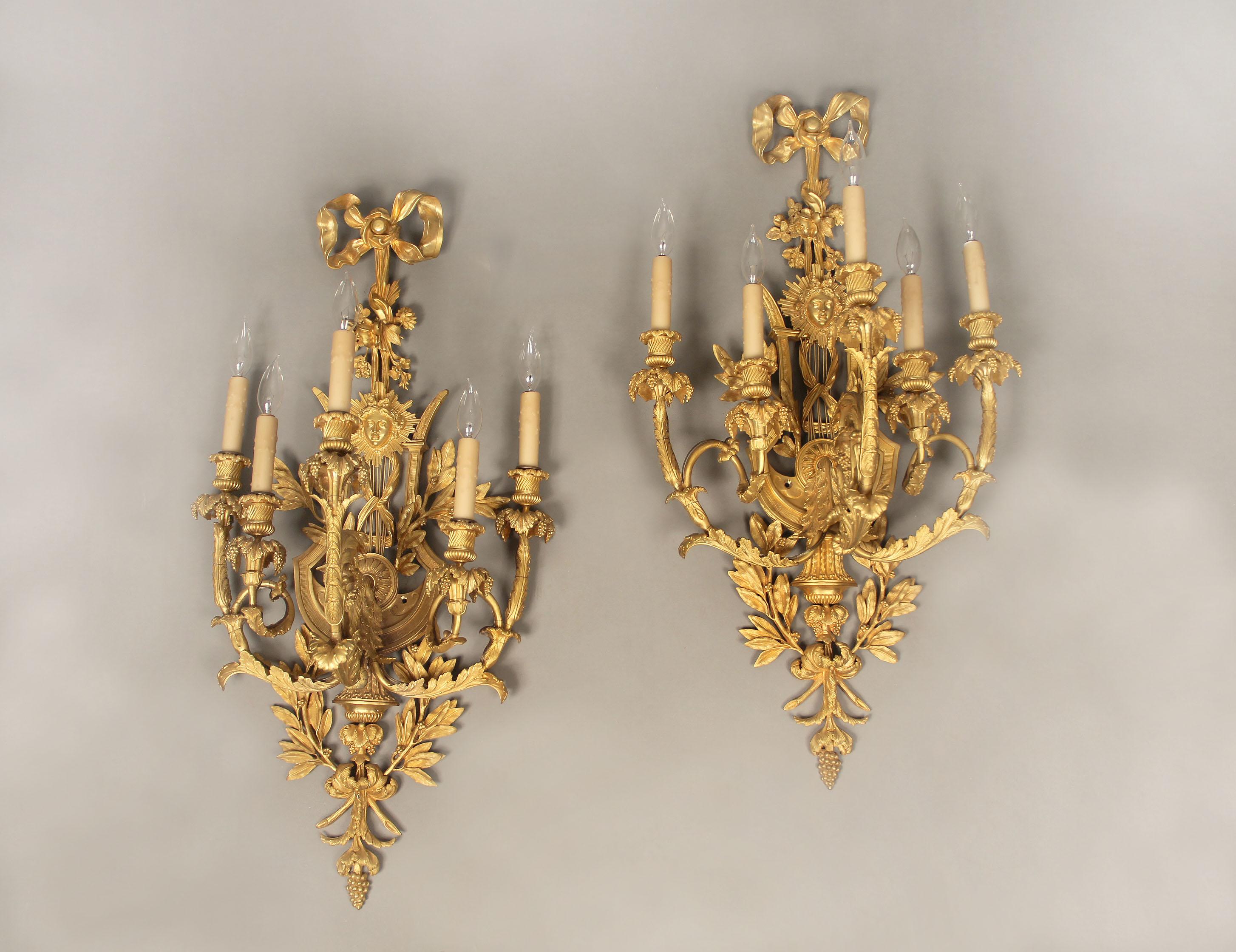 A large and elaborate pair of early 20th century gilt bronze five-light sconces

Each backplate shaped as a lyre, centered with a mask of Apollo, the top with a bow knotted ribbon, the arms and base decorated with leaves and berries.