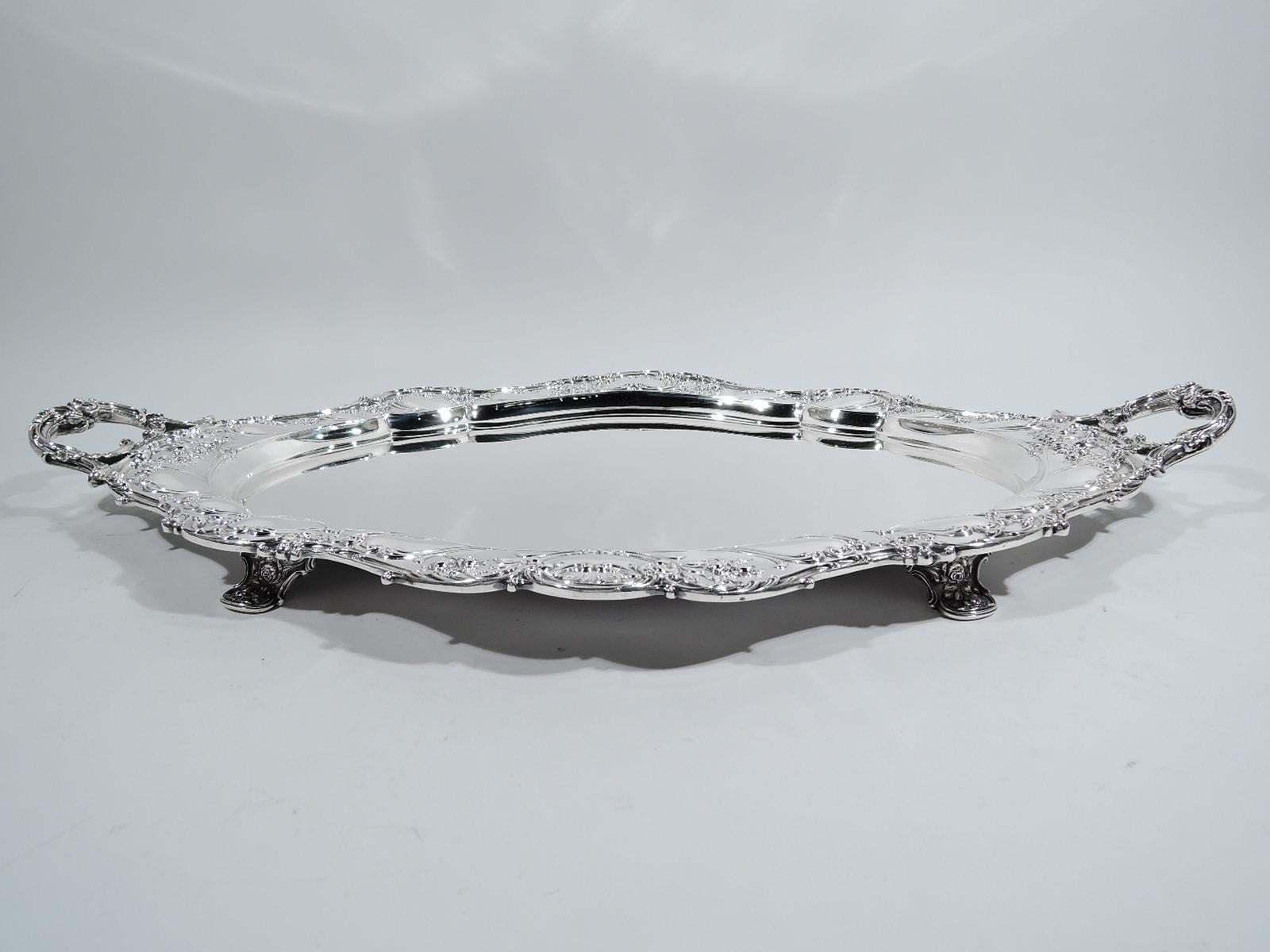 Turn-of-the-century sterling silver tea tray. Made by Gorham in Providence. Plain and shaped oval well. Scrolled rim and chased garlands, blossoms, and wave-like scrolled cartouches on shoulder. Open C-scroll end handles. Rests on four supports.