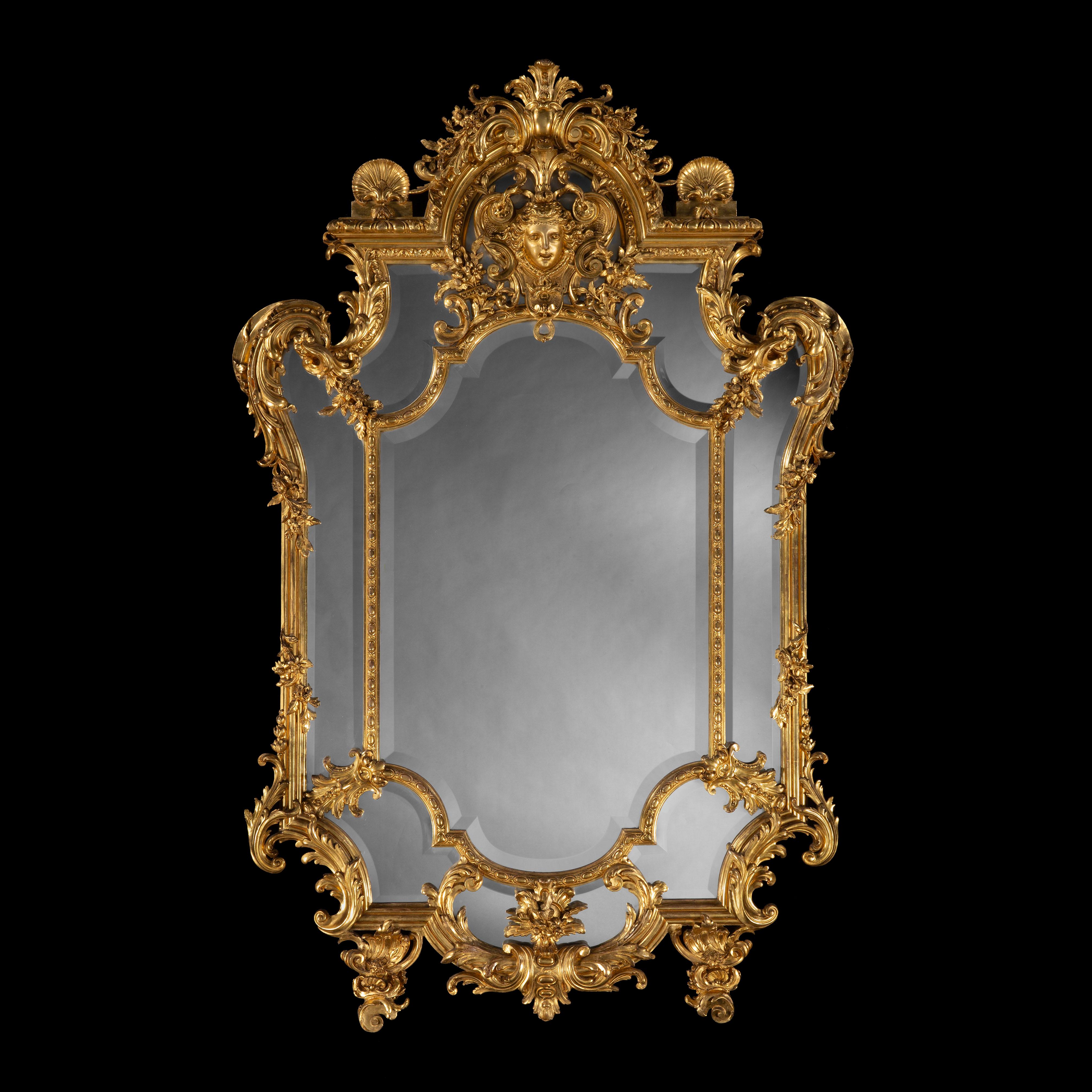 An Impressive Carved Giltwood Mirror
In the Régence Style

Of generous proportions, with effusive carved details, the central bevelled glass surrounded by an outer reserve of mirrored panels, the frame carved with scrolls, floral garland, pendants