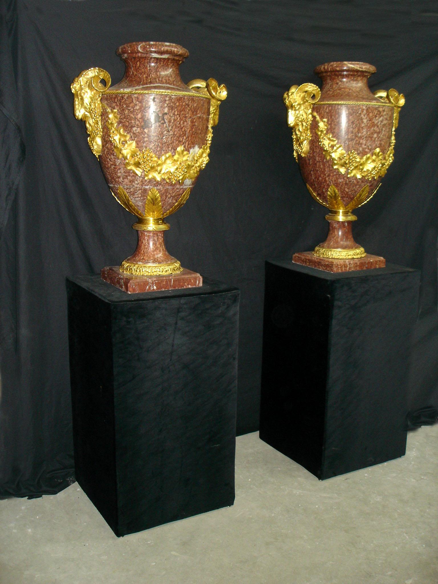 A large and impressive pair of late 19th century gilt bronze mounted rouge marble urns.

Each urn mounted with bronze ram heads connected by bronze wreaths.
