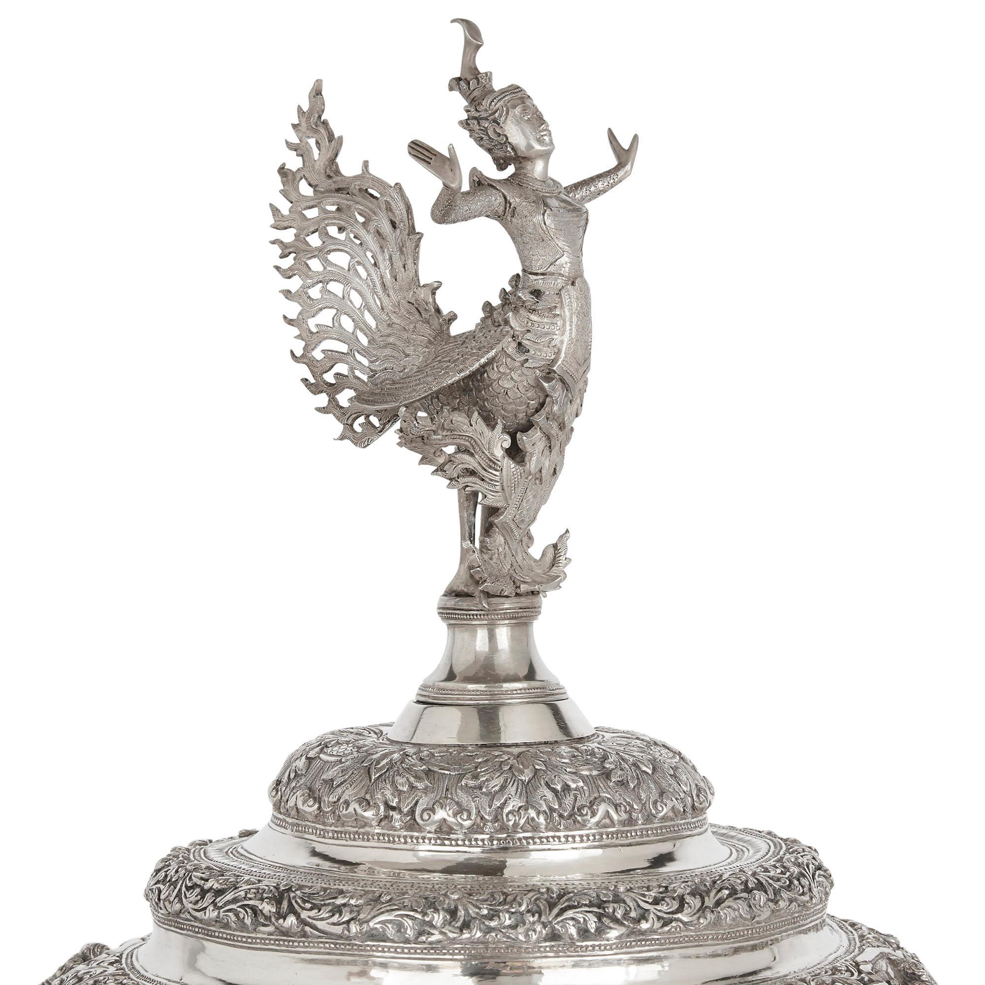 This large silver centrepiece vase is an aggrandised version of a type of Buddhist bowl from Burma (modern day Myanmar).

The surface of the bowl is divided into eight scenes from Burmese legend, the vignettes including warriors, courtiers, and