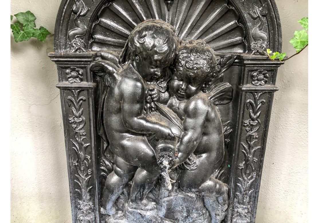 Heavy cast iron Putti Fountain for exterior or interior use. Black painted arch shaped top with high relief decoration of two putti pouring from a vessel as the fountain spout. Foliate decorated borders and a shell and floral crest. 
The shaped
