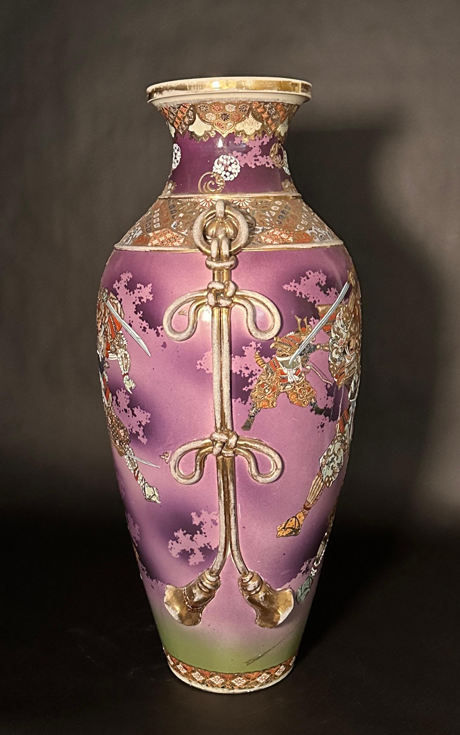 Grande e insolito vaso giapponese Satsuma con figure di samurai sul fronte e sul retro Fondo viola con colori dorati e tradizionali. Spalle intagliate e modellate ad anello e a fiocco, con Corde e nappe ai lati. Foro sul fondo.