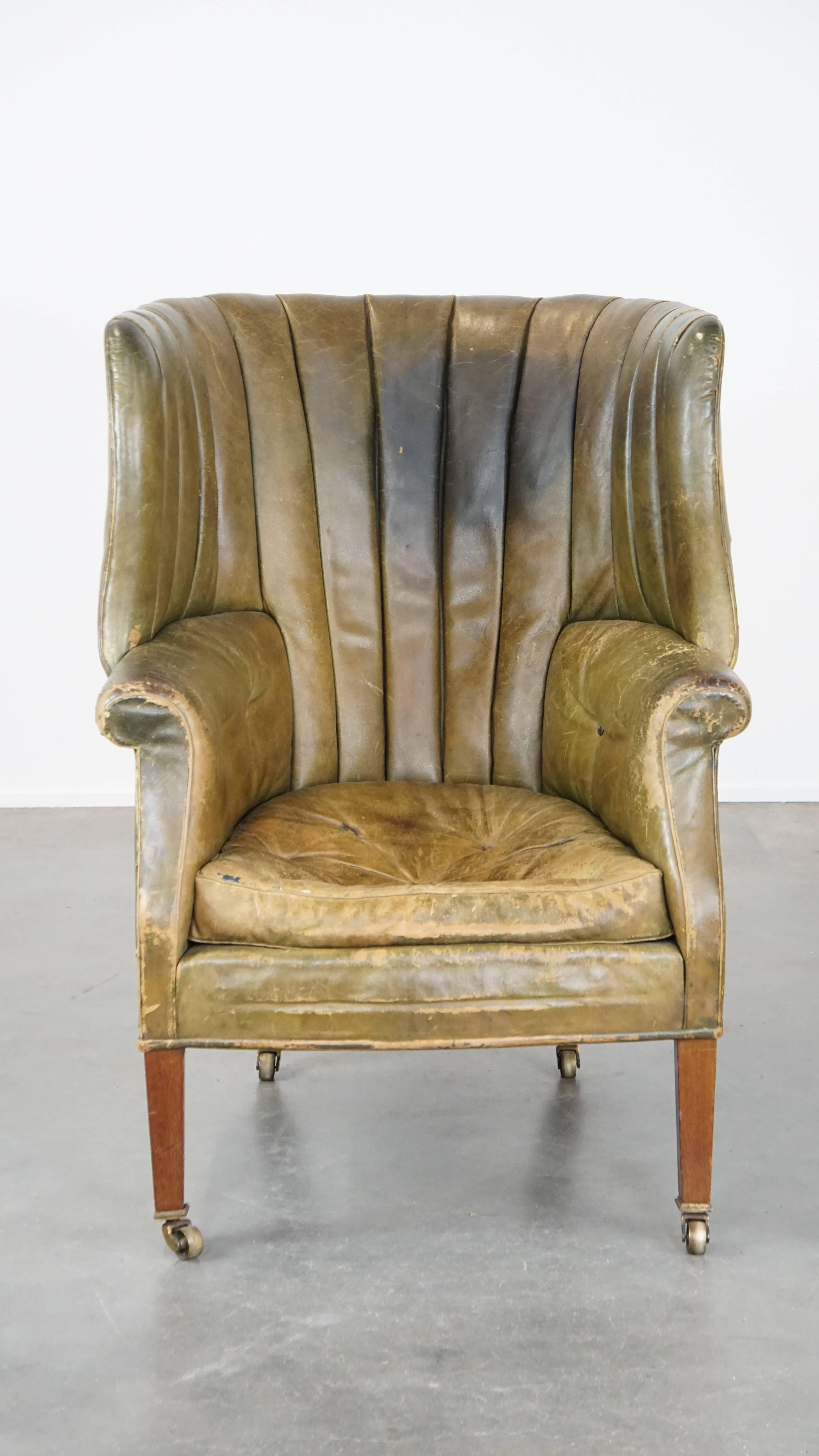 This amazing green leather barrel back wingback chair, known for its unique design and comfort, absolutely lives up to expectations. It has a beautifully lived-in character, with gorgeous colors and buttons on its seat and lower back. Its wheels