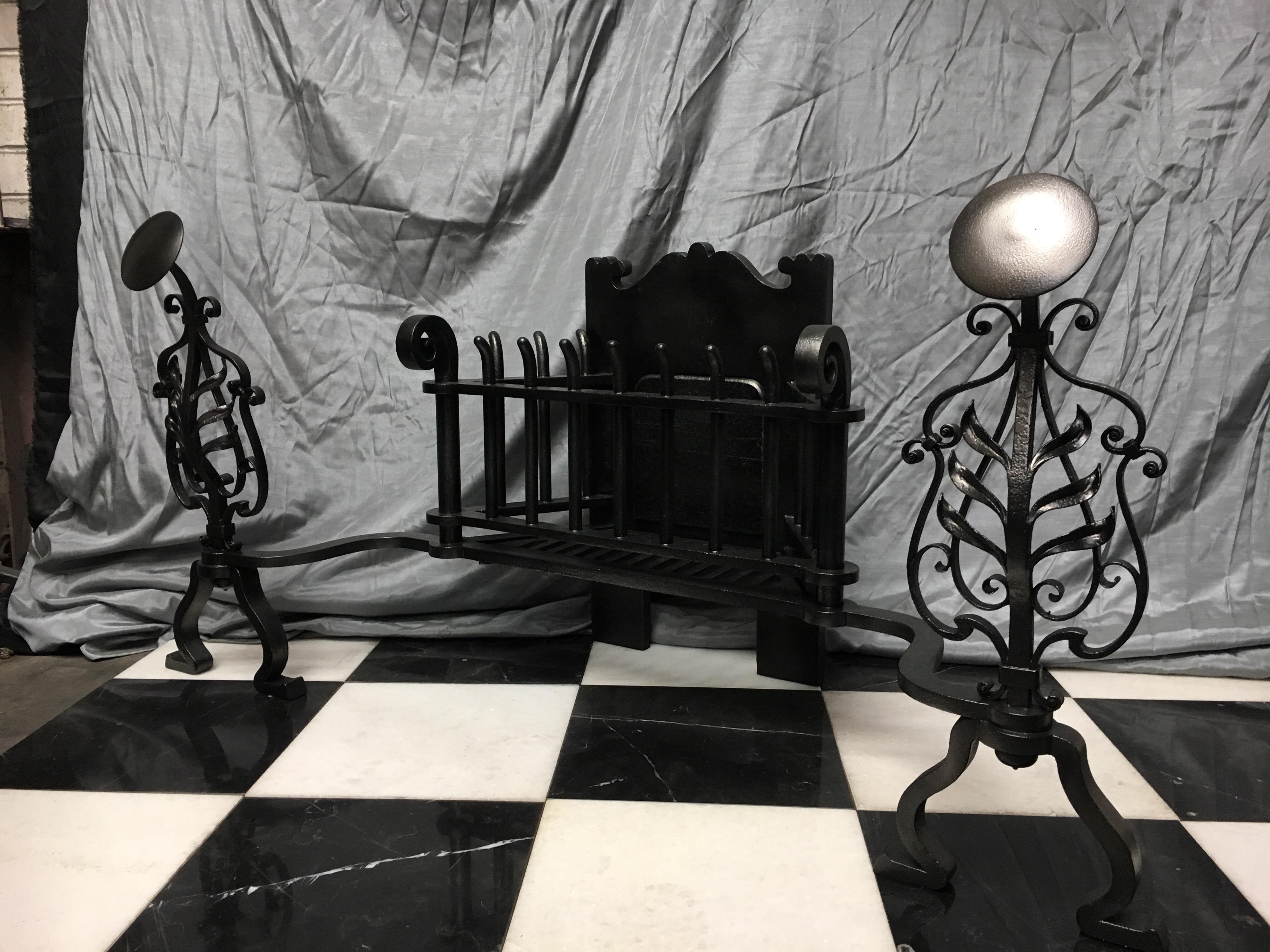 A large and substantial antique wrought iron fire grate in the Arts & Crafts style, a heavy set cartouche back plate supporting vertical fire bars flanked by round standards displaying Pig tail finials. Flanked in turn by tall ornate foliage and