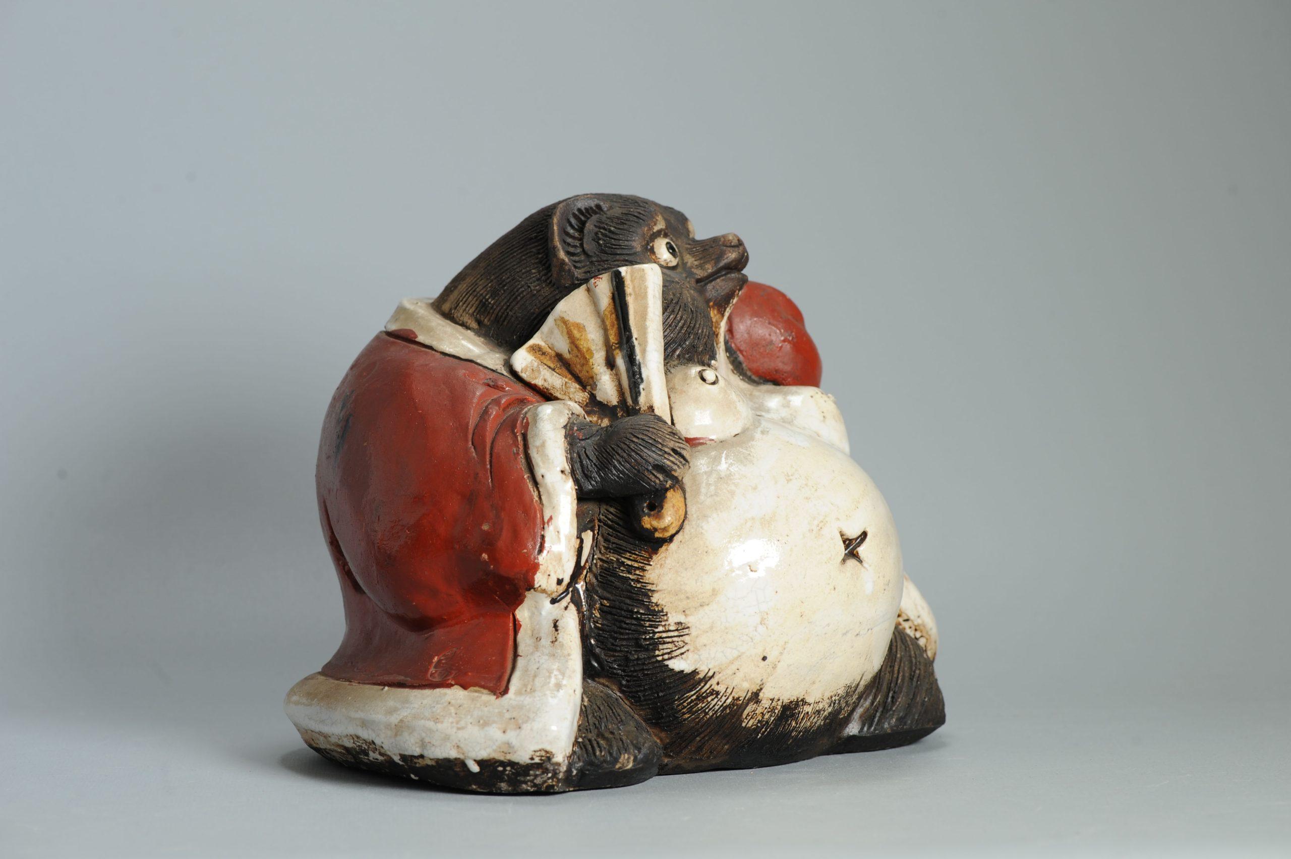 Cette figurine en céramique représente un tanuki japonais (狸 - chien viverrin), une créature bien connue du folklore japonais. Ces figures sont considérées comme des symboles de bonne fortune, de prospérité et d'espièglerie. Elles sont souvent