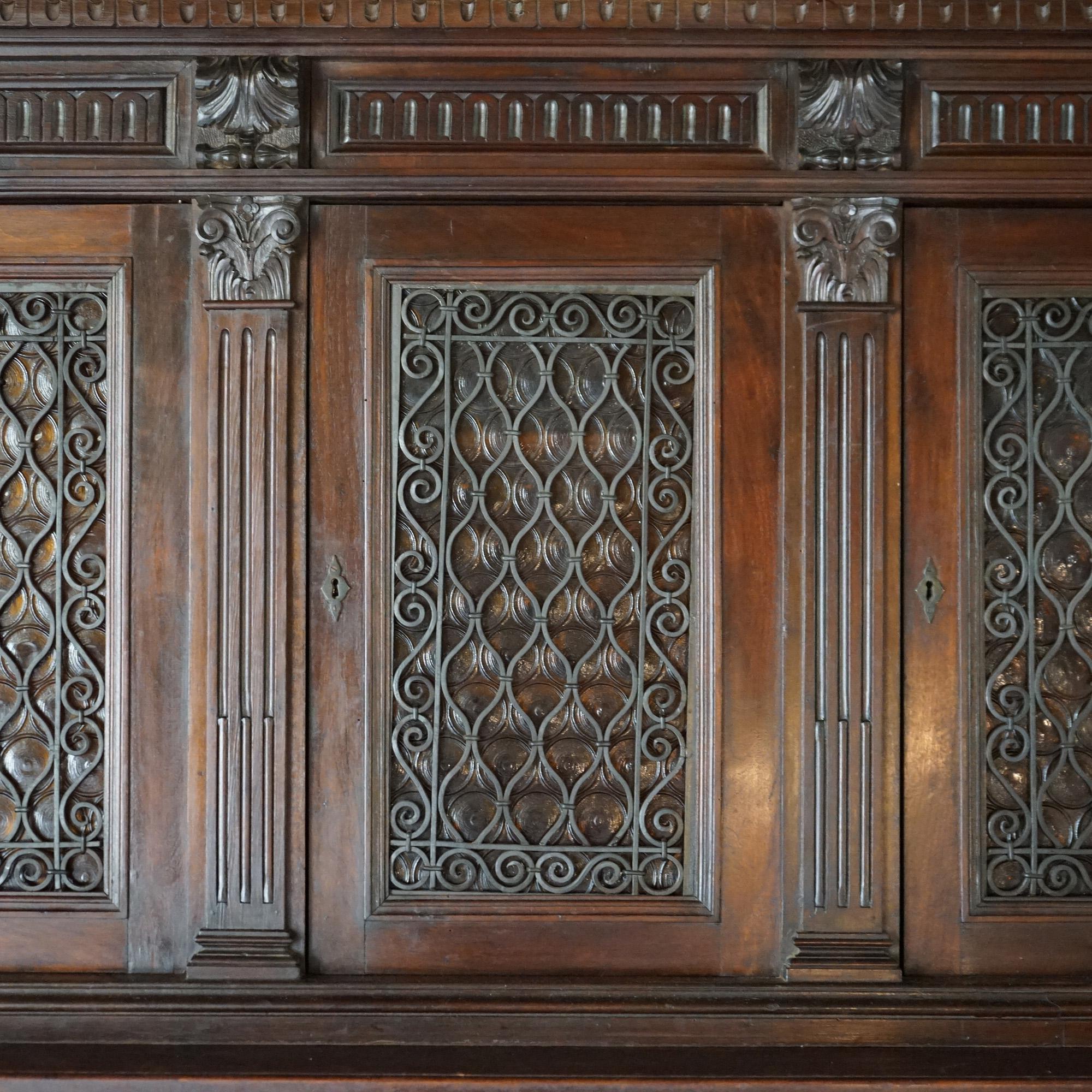 An antique Baroque hunt board offers walnut construction with upper case having three bottle glass doors with scrolled wrought iron grates and opening to divided and shelved interior; lower case with three upper drawers over thee blind cabinets,