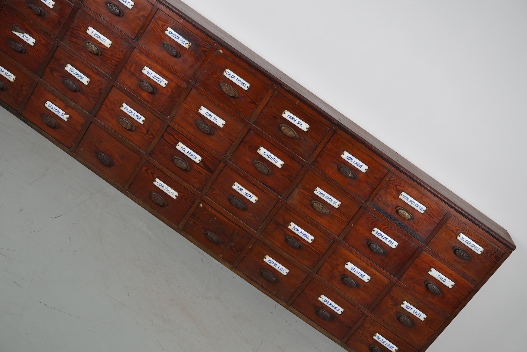 This apothecary bank of drawers was designed and made circa 1900 in Belgium. It was made from pitch pine with cast iron art nouveau style handles and enamel name plates for the contents. The cabinet has retained a rich patina with some natural