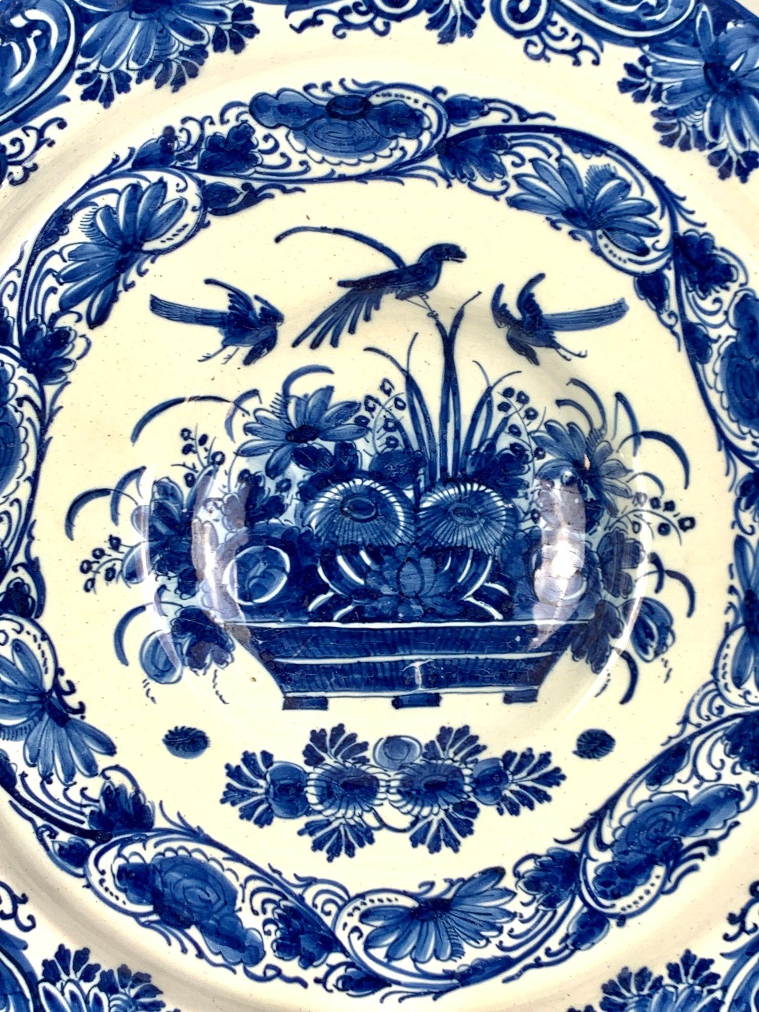 Questo caricatore Delft olandese blu e bianco è stato dipinto a mano in un intenso blu cobalto nel XVIII secolo, intorno al 1780.
La scena centrale presenta una coppia di uccelli in volo sopra un cesto pieno di fiori e steli di foglie.
Gli uccelli -