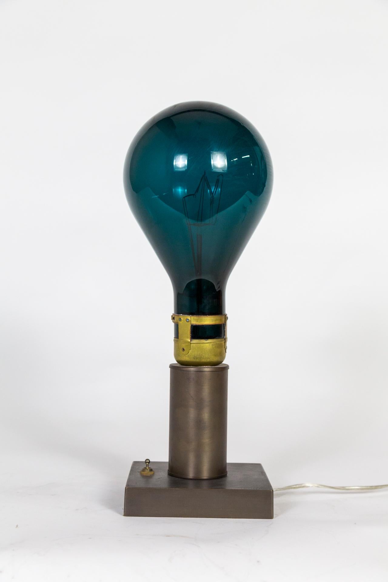 A table lamp made from a giant, vintage light bulb in blue-turquoise; illuminated from within by an Emery Allen LED e12, (5 watt). Finely finished with a vintage switch and square, brass base tinted in antique tone. Measures: 17.25