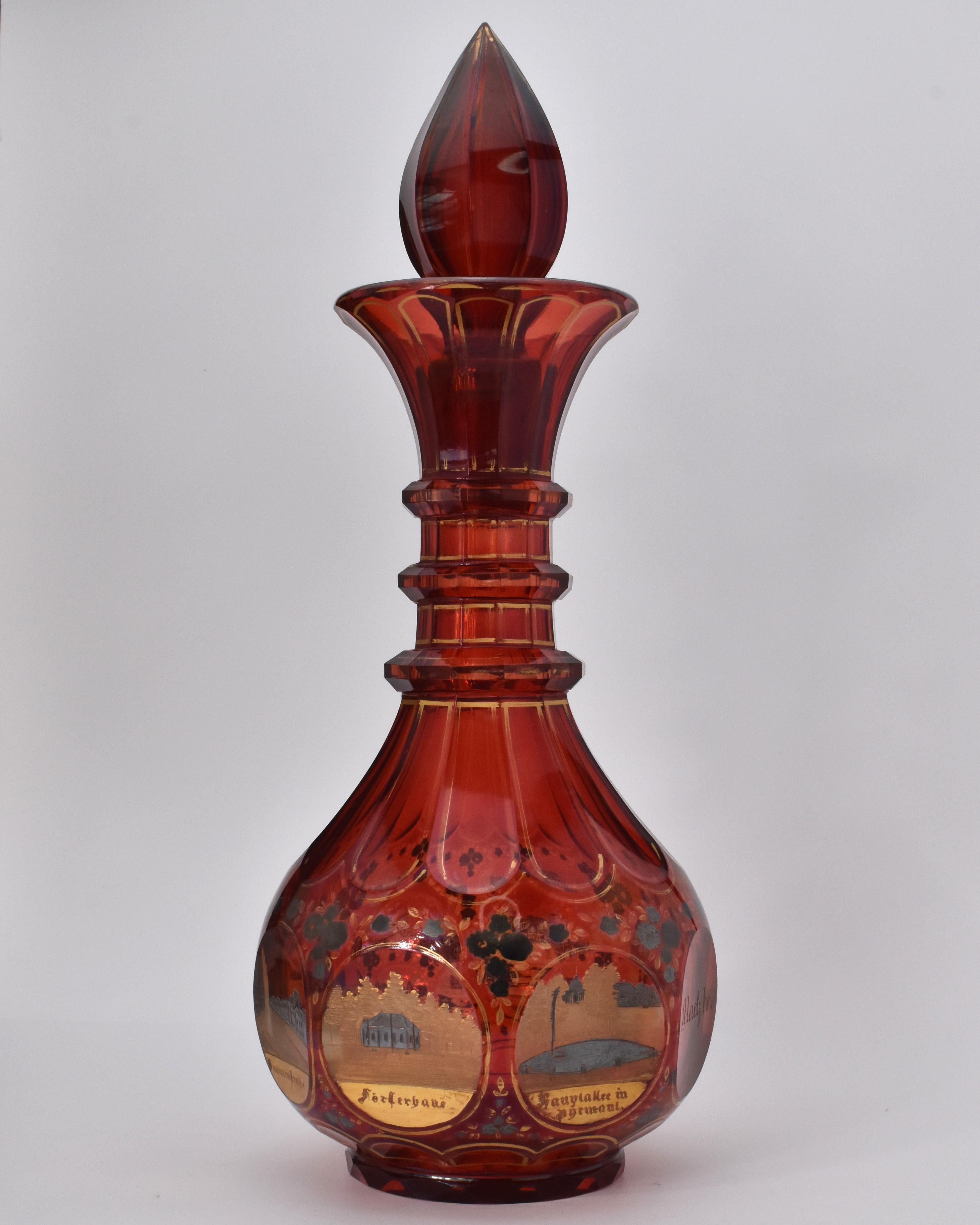 grande carafe avec bouchon en verre transparent rouge rubis profond

décoré sur tout son pourtour de multiples scènes gravées, de rehauts d'émail argenté et de dorures

le col est appliqué avec trois anneaux

Bohemia, 1840
La carafe a une hauteur de