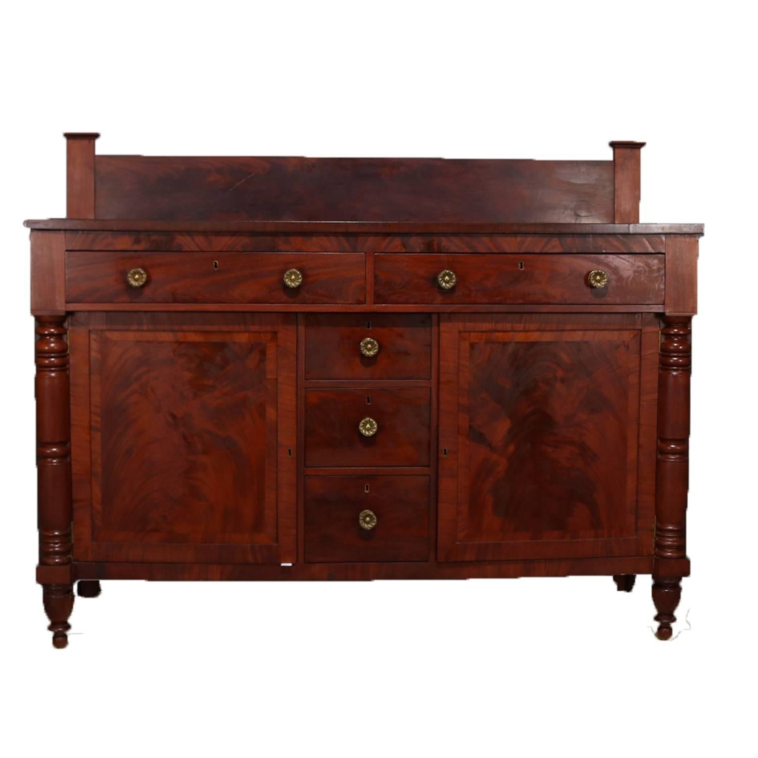 Antique classical American Empire sideboard features banded flame mahogany case having back splash and two upper long drawers over three stacked smaller drawers flanked by cabinets with shelved interiors, seated on turned turnip legs, cast bronze