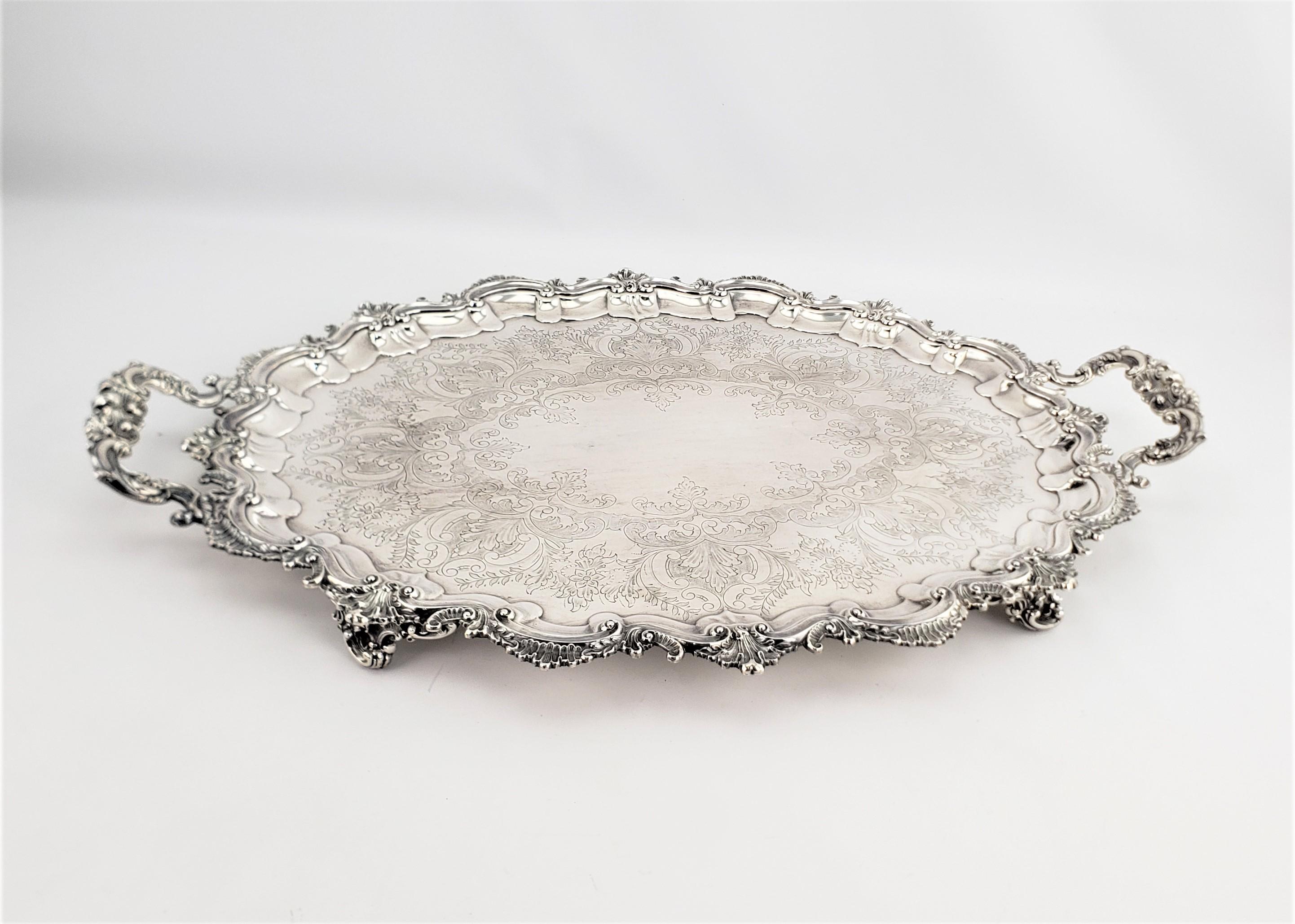 This large footed silver plated serving tray is marked by an unknown maker, and originates from England and dates to approximately 1920 and done in a Victorian style. This large oval serving tray is done with an elaborate floral decoration along the