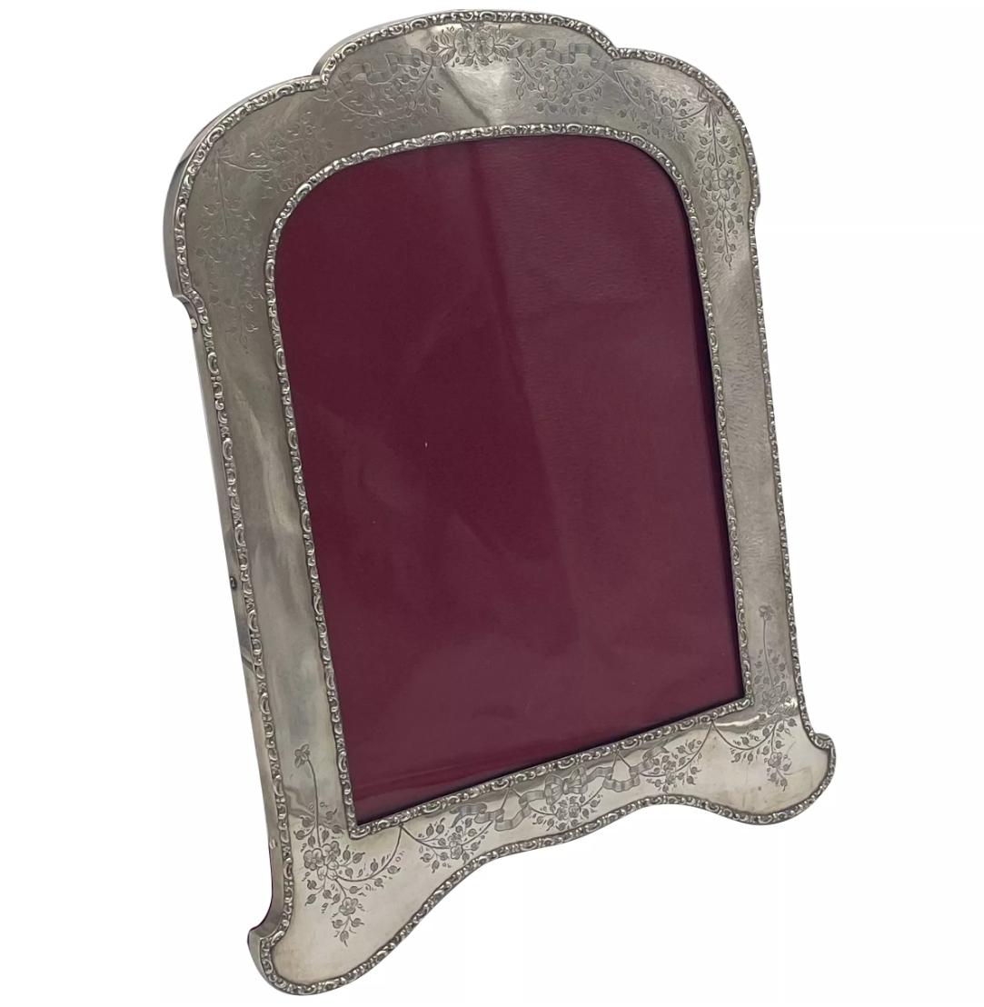 A large sterling silver photograph frame, made in England and hallmarked In Birmingham 1906.
The frame is beautifully engraved with ribbons and garlands of flowers, and will hold a photo measuring 28cm x 21.3cm. The frame is mounted on a red velvet