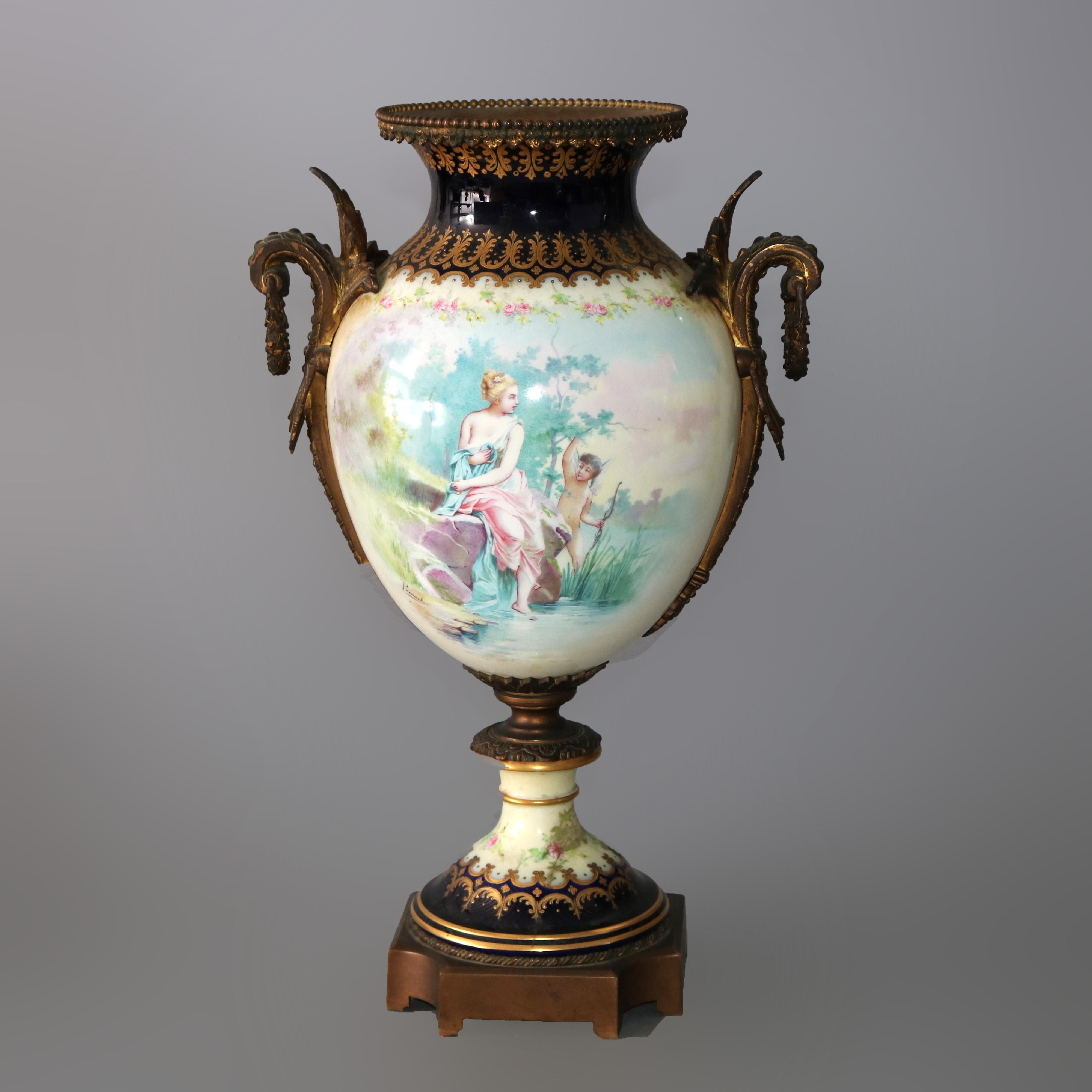 An oversized antique French urn attributed to Sevres offers porcelain construction with an artist signed hand painted reserves by Pesaud depicting a woman and Cupid in countryside setting on vessel with cobalt blue banding, foliate cast bronze