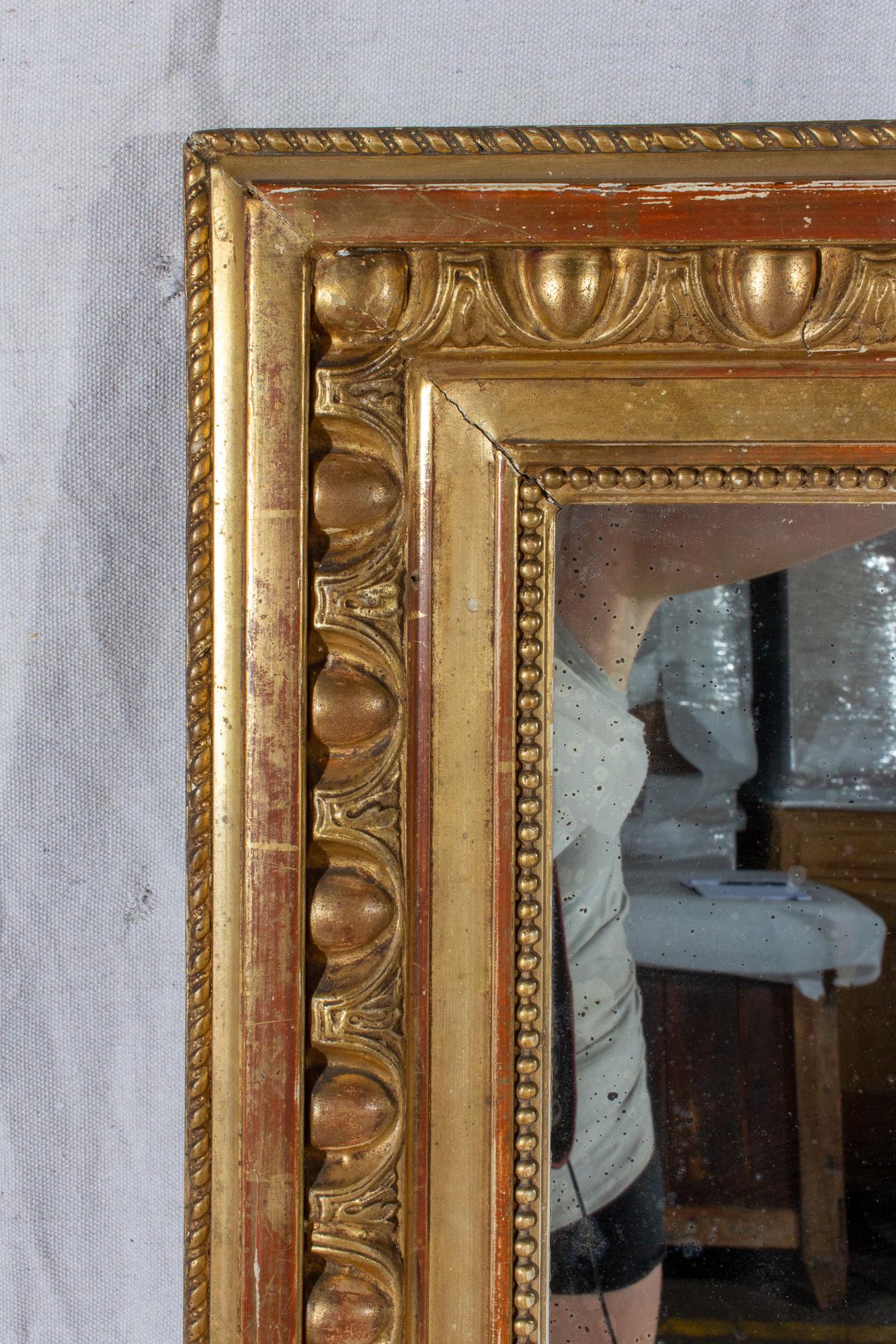 This is an antique French mirror with original glass, in a decorated, rectangular gilt frame featuring gold, bronze and red tones. The frame is somewhat distressed, with losses to the plaster and gilt. The mirror itself does show some age and there