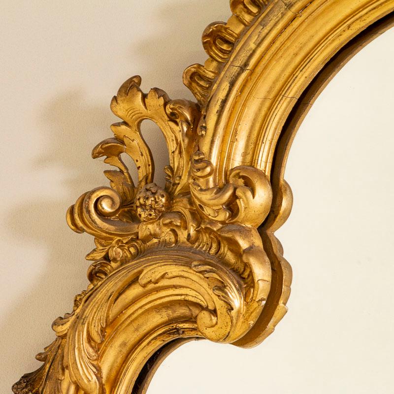This stunning gold gilt mirror is almost 7' tall and is crowned with carved flowers and flourishes. The sides and lower corners are accented with elegant effect. There is expected age-related wear as seen where the gilt has been worn off, chipped or