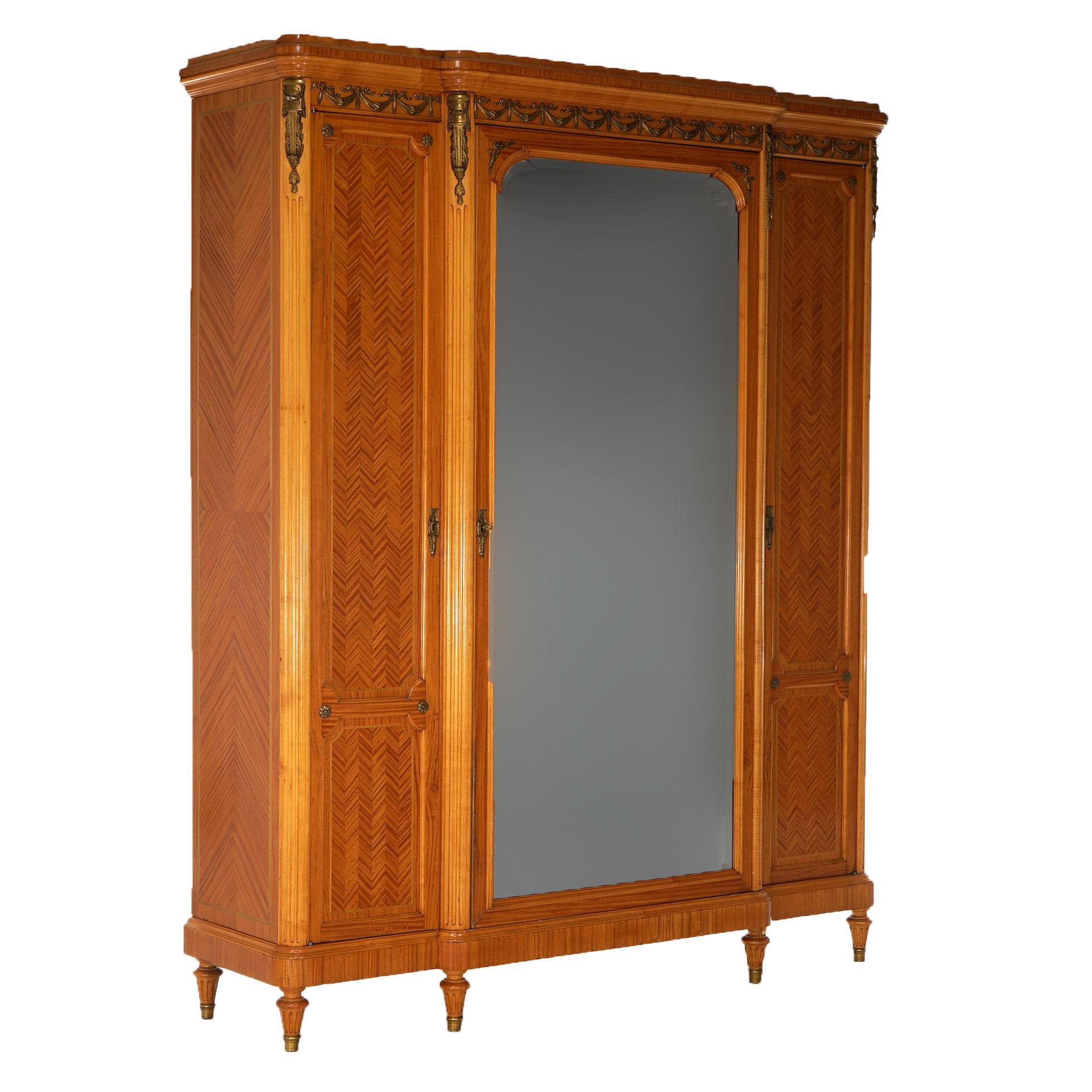 An oversized antique French Louis XIV style armoire offers satinwood construction with chevron style inlaid panels, foliate garland cast ormolu mounts, mirrored door opening to shelved interior and flanked by blind cabinets, and raised on Marseille