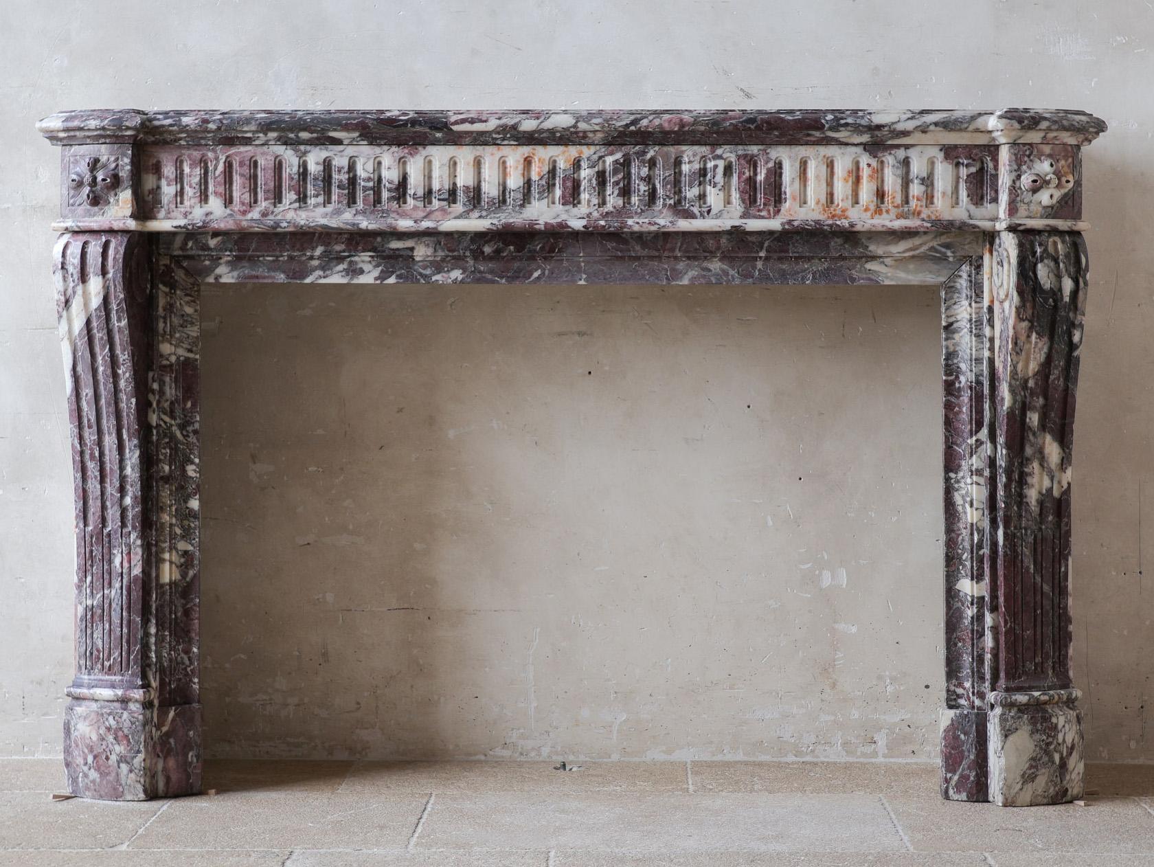 This Large Louis XVI fireplace features a convex fluted fronton adorned with rosettes at the corners. The fluted jambs have an elegant pied galbé shape and are decorated with graceful scrolls on the sides. Crafted from exceptionally beautiful Fleur
