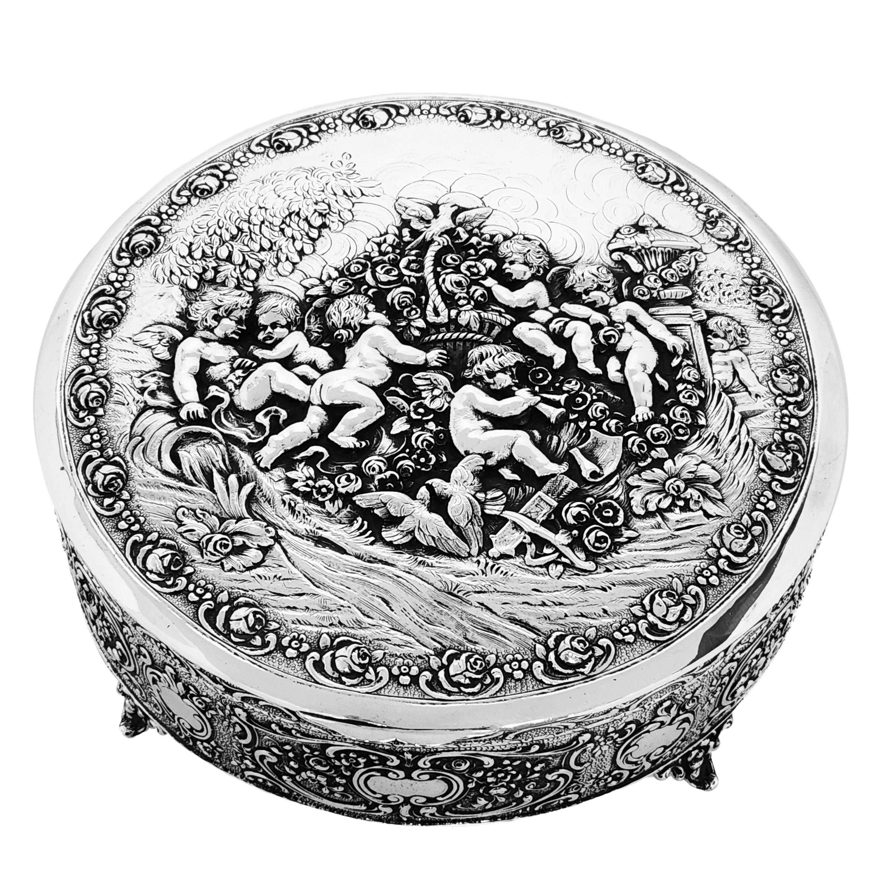An impressive Antique Solid Silver Box with a round body and a fitted hinged lid. The Box is embellished on both the lid and sides with ornate and detailed chased designs. The Lid shows a group of cherubs or putti frolicking in a floral garden,
