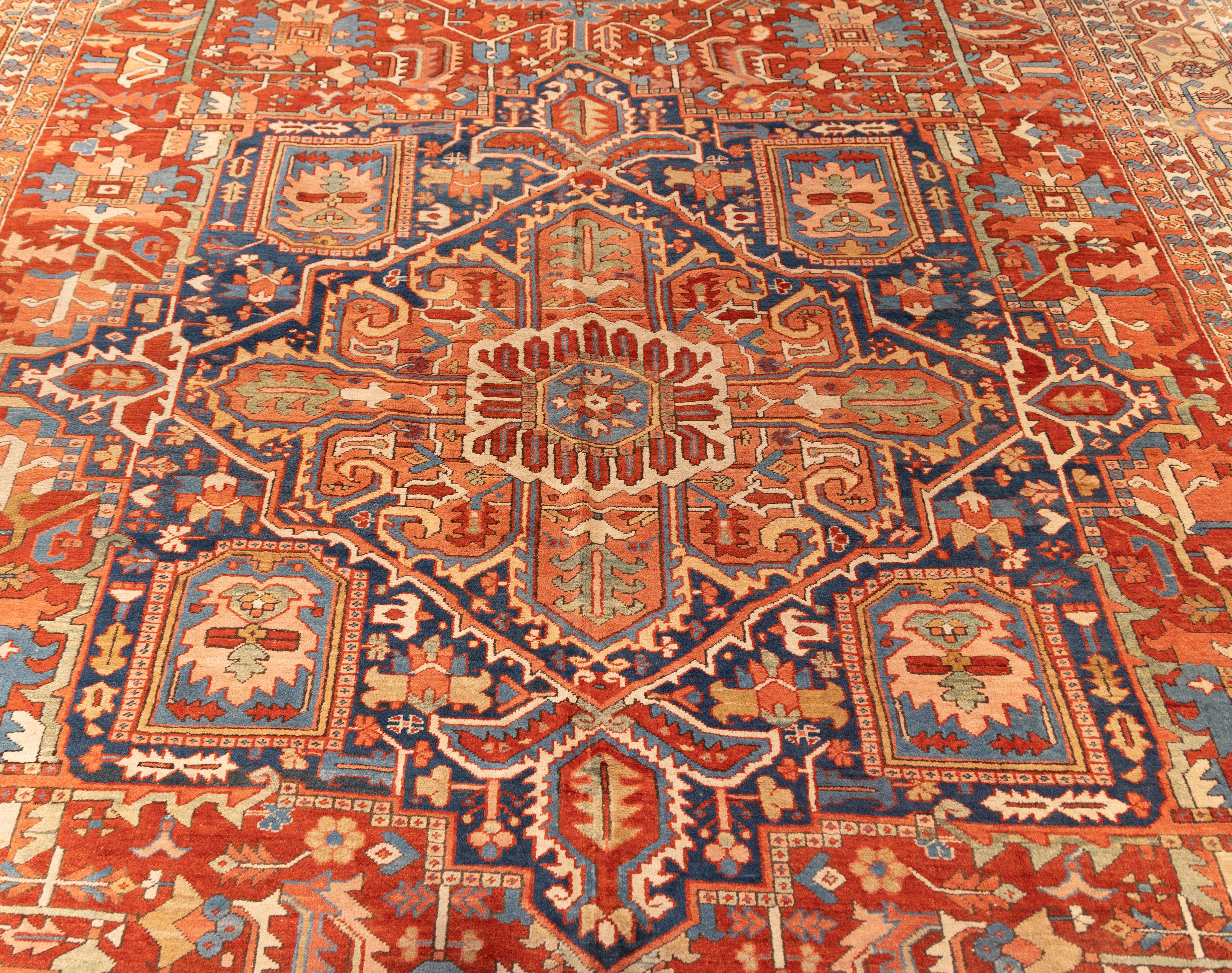 Oversized Early 20th century Heriz featuring a bold central medallion in deep navy, set against a vibrant rust-red field. Angular floral motifs, stylized rosettes, and stepped medallion corners are framed by intricate ivory spandrels and a