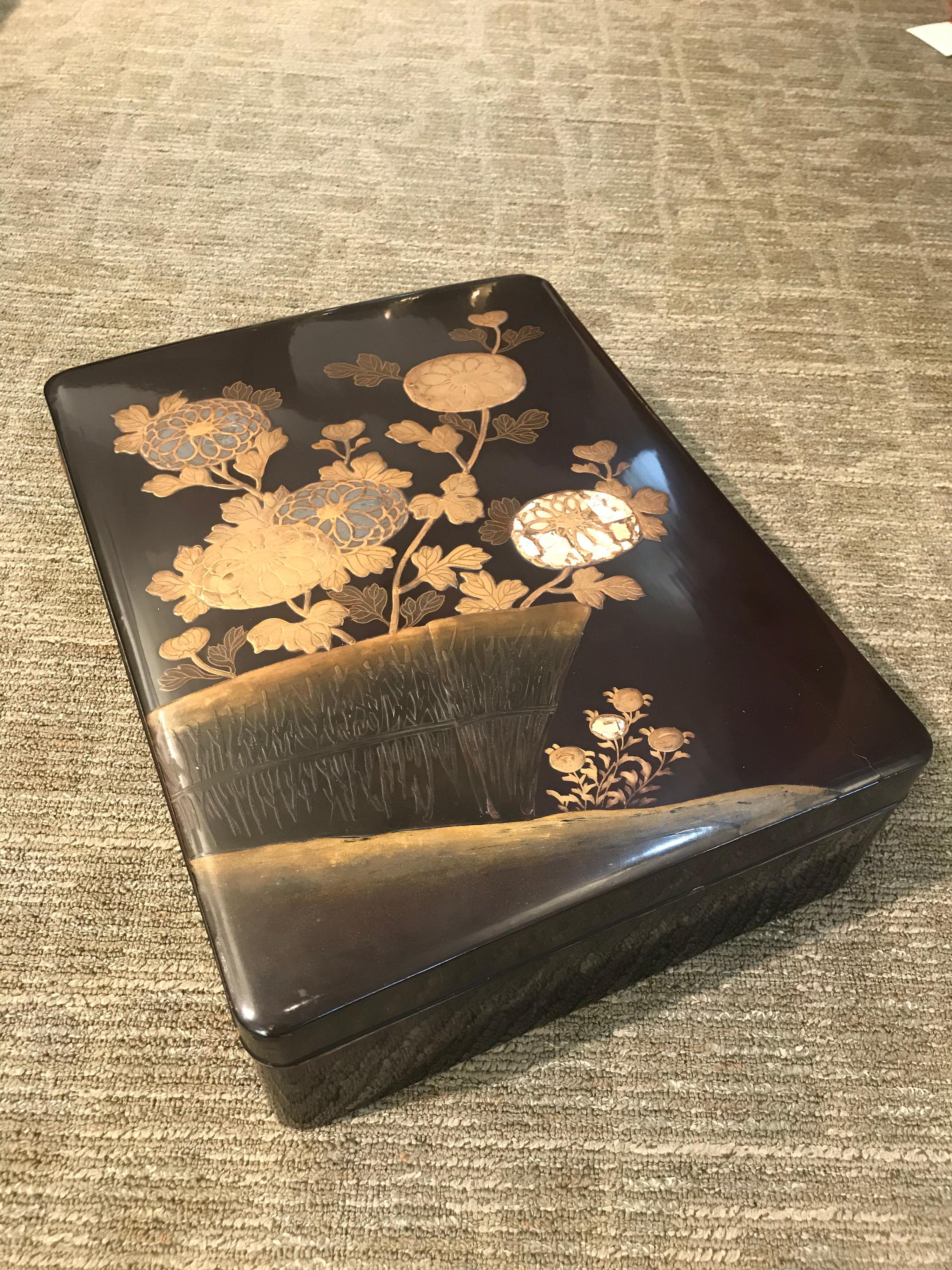 A large and beautifully decorated antique Japanese lacquer box.

Finished in very dark brown lacquer, the box is decorated with raised lacquer and mother of pearl design of flowers and a reed fence.

Inside is finished in speckled gold lacquer,