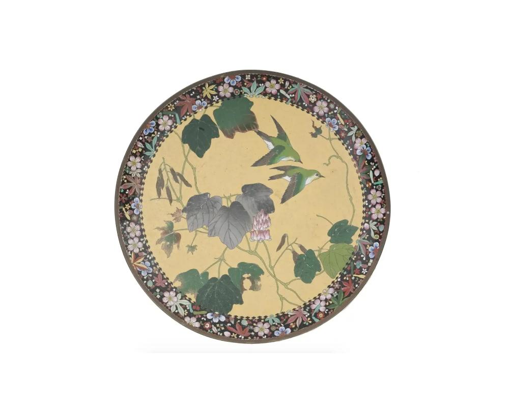 A large antique Japanese Meiji period cloisonne enamel metal charger plate depicting a multicolored picture of two birds hovering over graceful acacia branches on a pale yellow ground. Features a highly detailed polychrome border depicting cherry