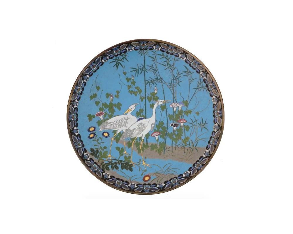 An extra large antique Japanese Meiji era enamel over brass charger plate. The plate is enameled with a polychrome image of naturalistic cranes in blossoming flowers and trees on blue ground made in the Cloisonne technique. The border is decorated