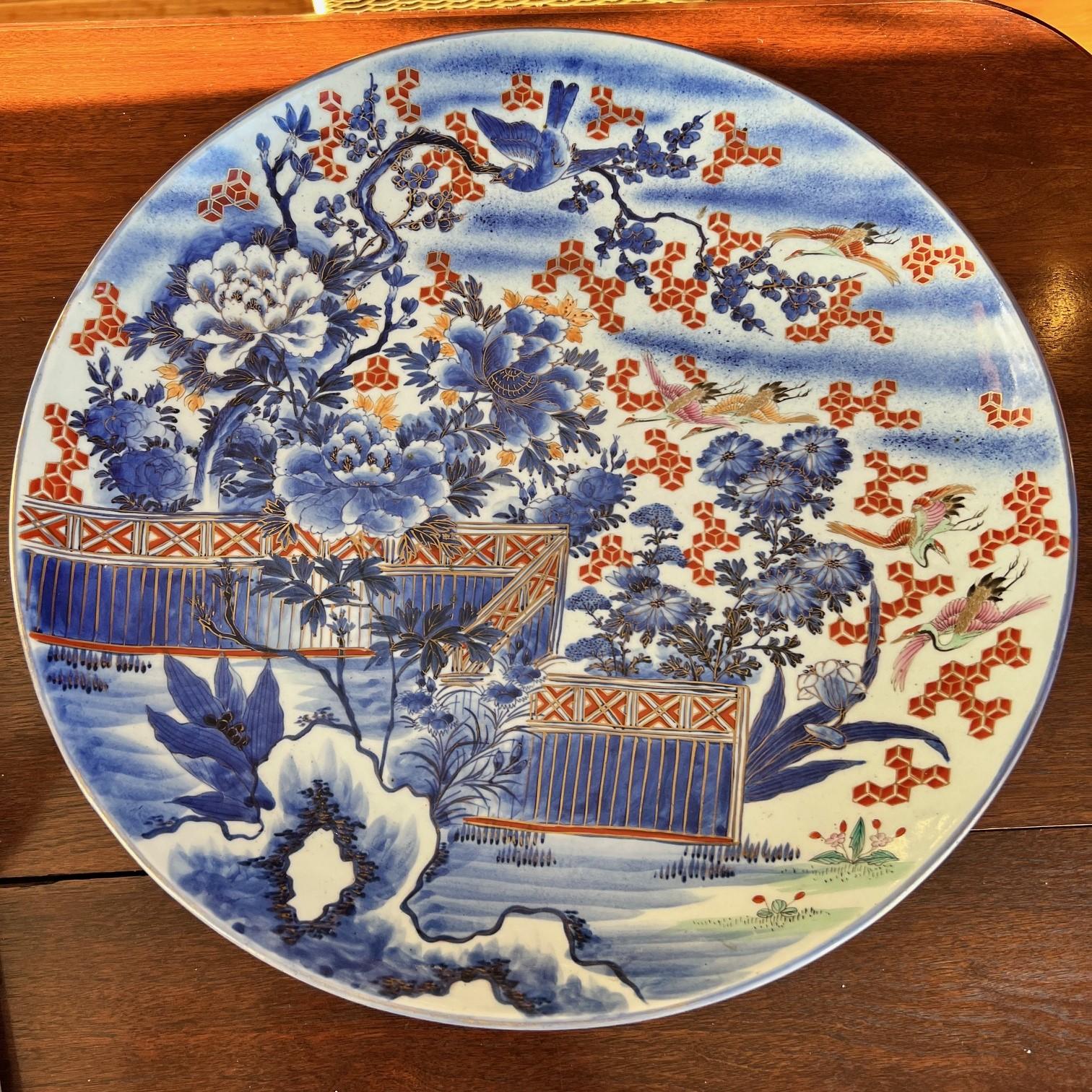 Late 19th C. large, decorative Japanese Imari plate featuring intricate hand-painted designs in blue, red, and gold. The central motif includes blue floral patterns with accents of red geometric shapes and cranes in flight, symbolizing longevity and