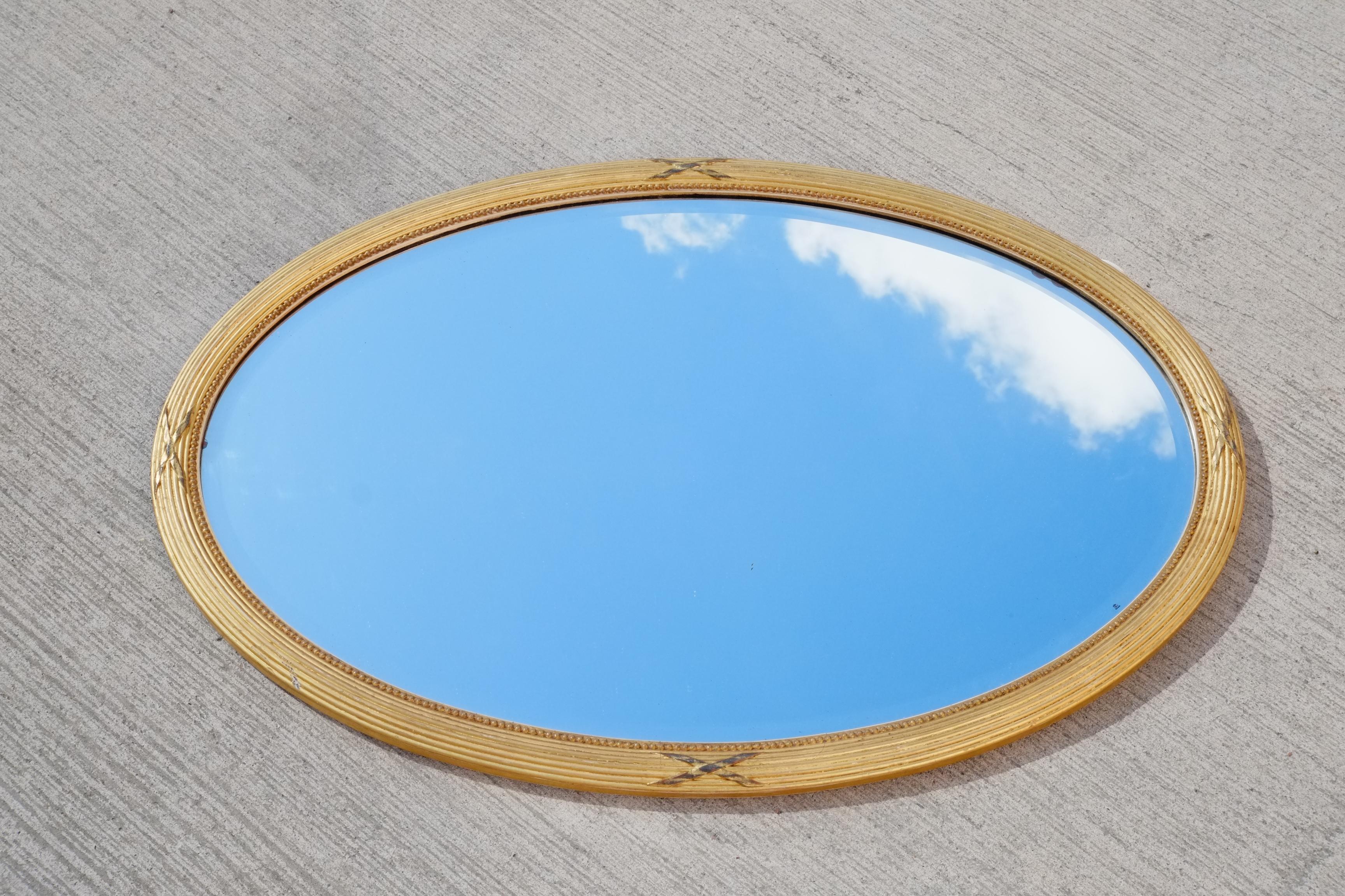 A beautiful large early 20th century gilt wood mirror. The frame is a large oval with reeded and X shaped detailing. A very soft gold colour which has a modest and elegant feel. Deep bevelled edge to the mirrored glass. This piece would look amazing