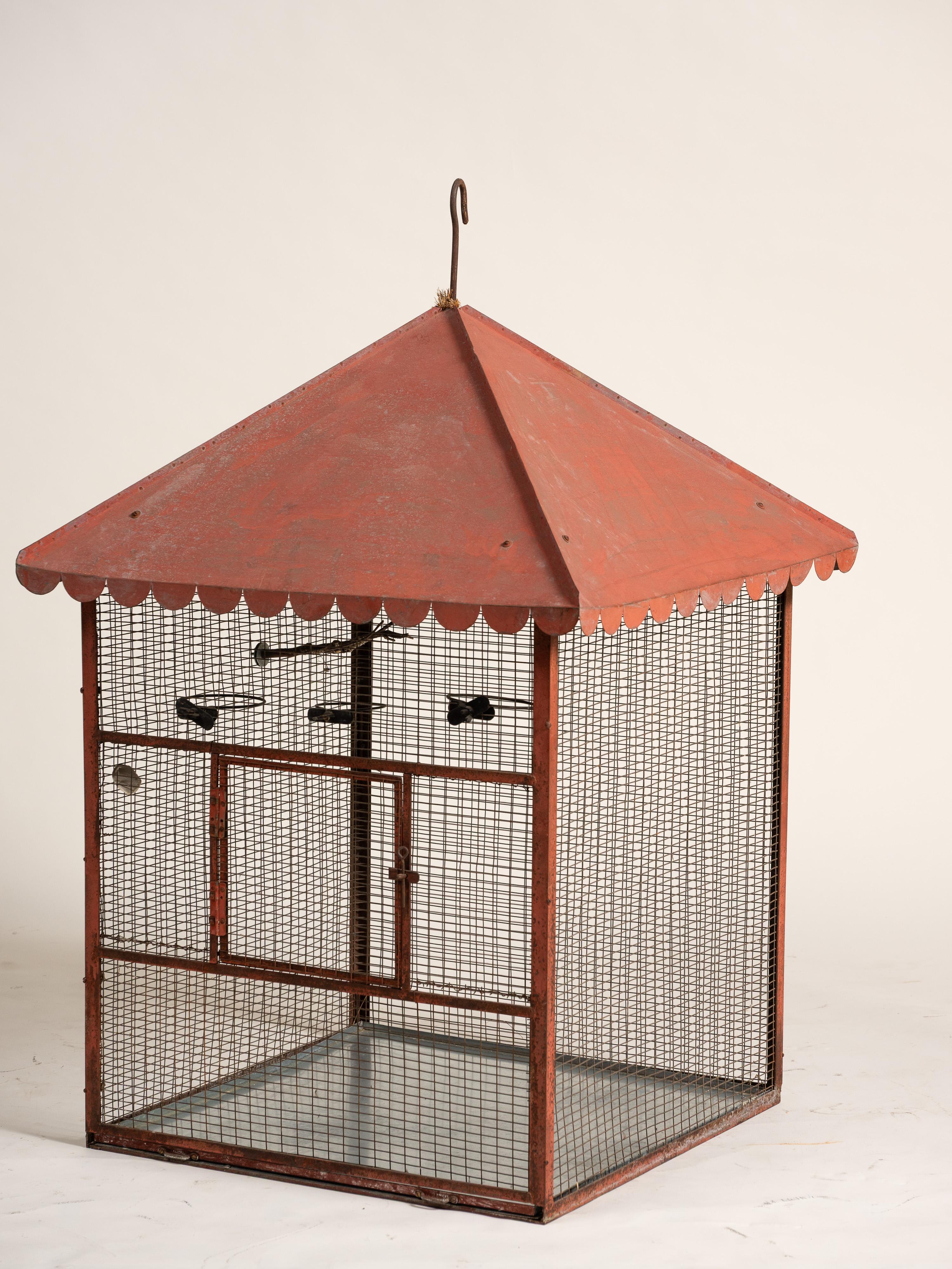 Cage à oiseaux vintage de style cottage ou farmhouse, en grillage rouge et à cadre métallique, avec une finition vieillie. La cage présente un toit rouge hexagonal de style pagode avec des bords festonnés et un crochet de suspension intégré sur le