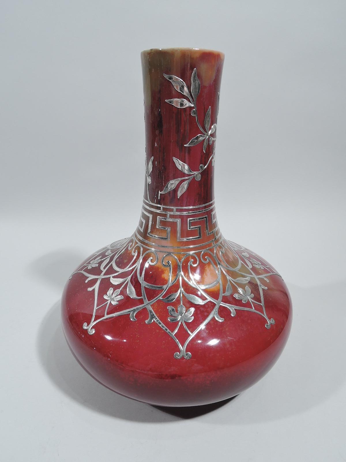 Large vase with engraved overlay, ca 1910. Wide bellied bowl and tall upward tapering cylindrical neck. Overlay in form of overlapping scrolled frames inset with stem flowers, and leafing branches and fretwork border. Red flambé ground with