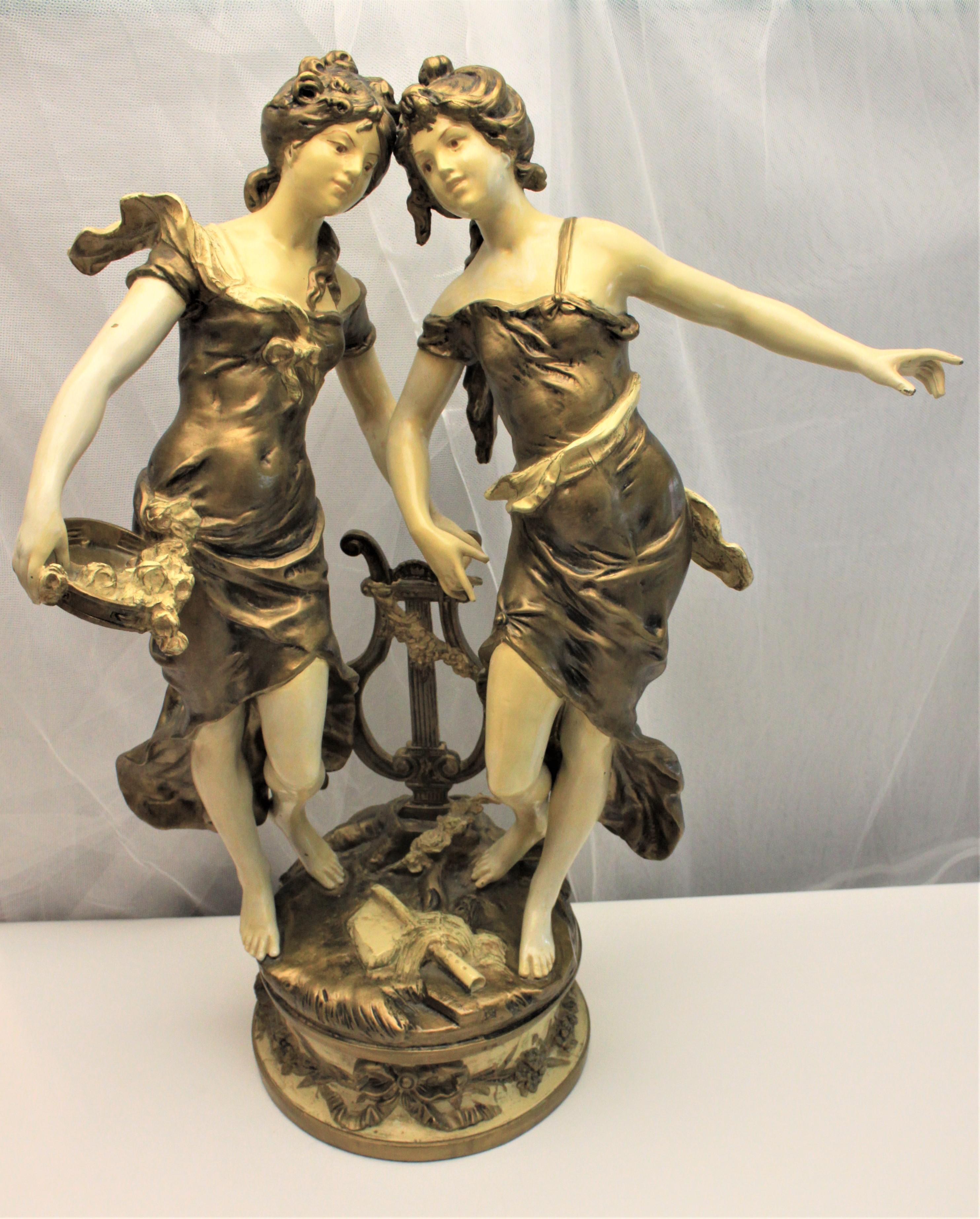 This large antique cast spelter and cold painted sculpture portrays two dancing women around a lyre. The sculpture is painted predominately in an off white or cream and olive green and the casting is very ornate and detailed. The sculpture is
