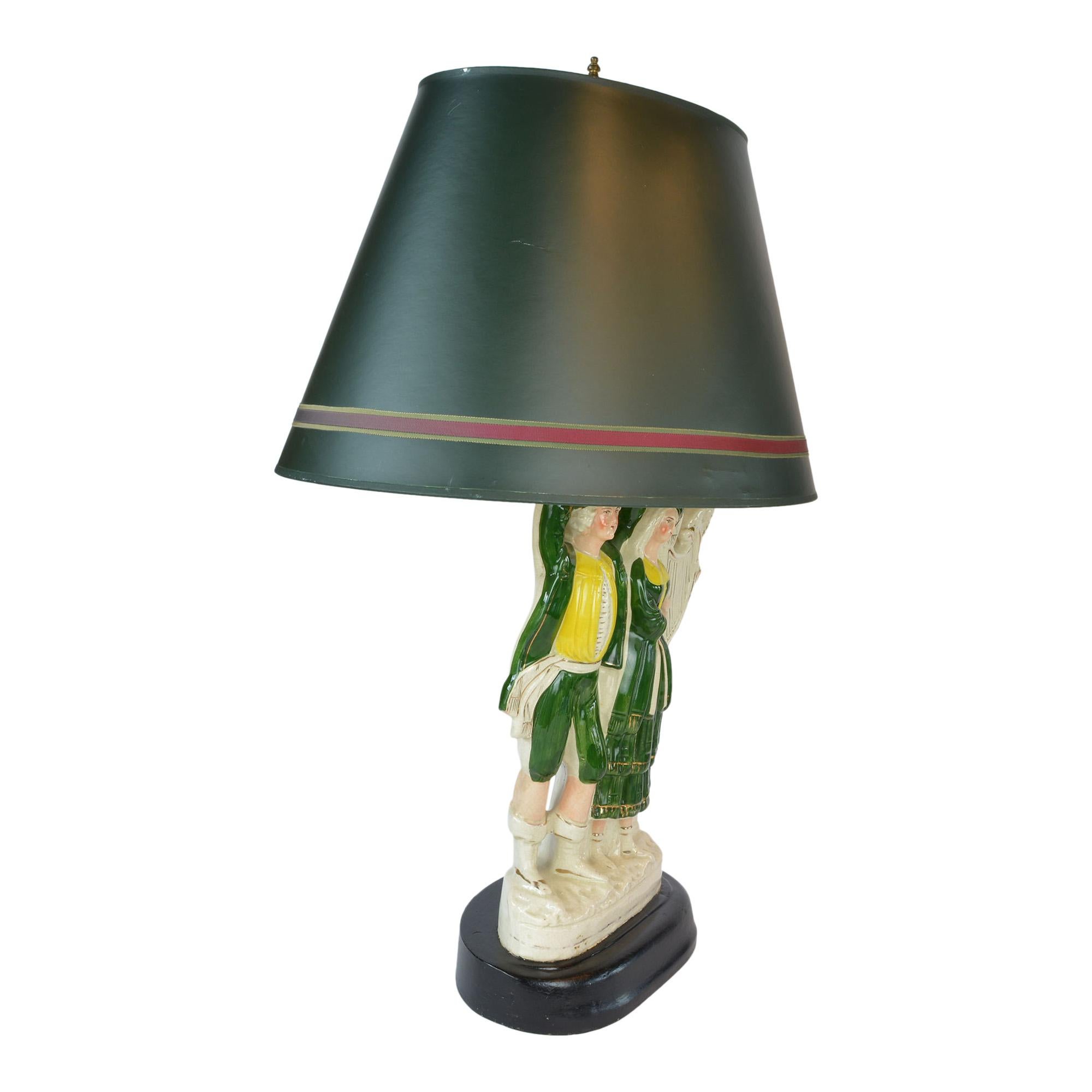 Large Antique Staffordshire figural has been converted into a lamp with an oval hard frame shade. The couple stands proud and united on the lamp each wearing green, yellow and white traditional attire. She is playing a harp accented in gold.
