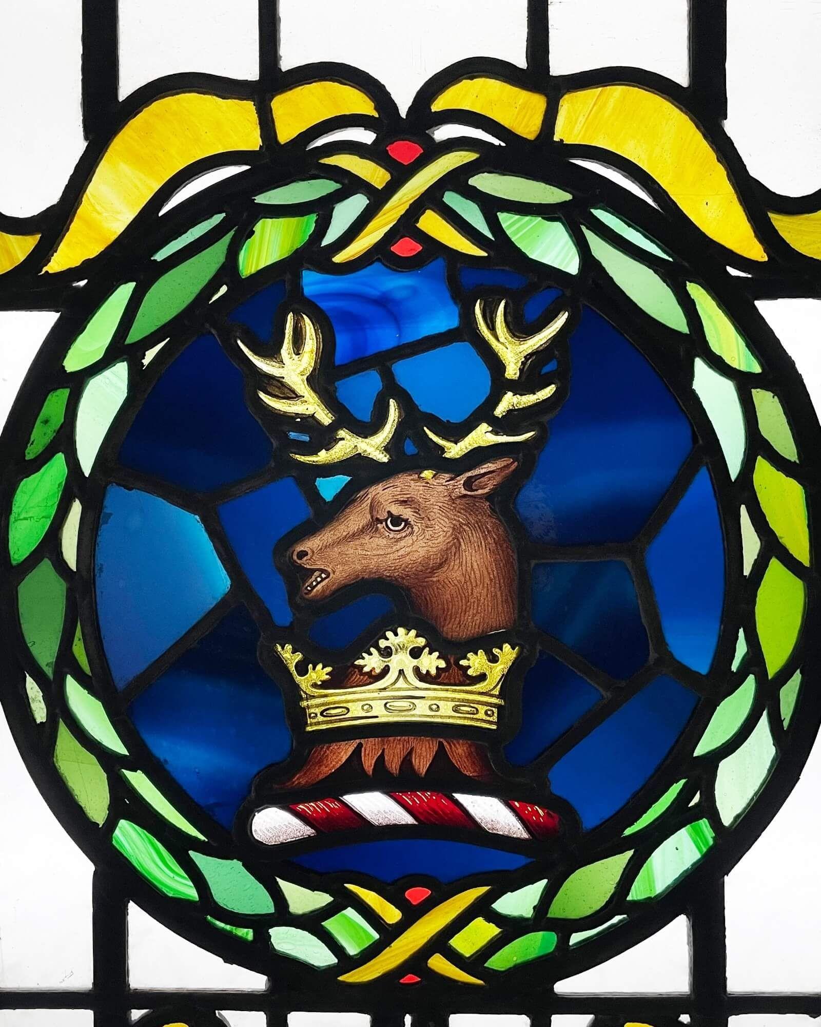 A large antique stained glass window of a crest depicting a stag in a wreath. Dating to circa 1900, this large panel exhibits vibrant handprinted colours. Pale green oval domes sit on top of the clear textured background.

The vivid imagery of this