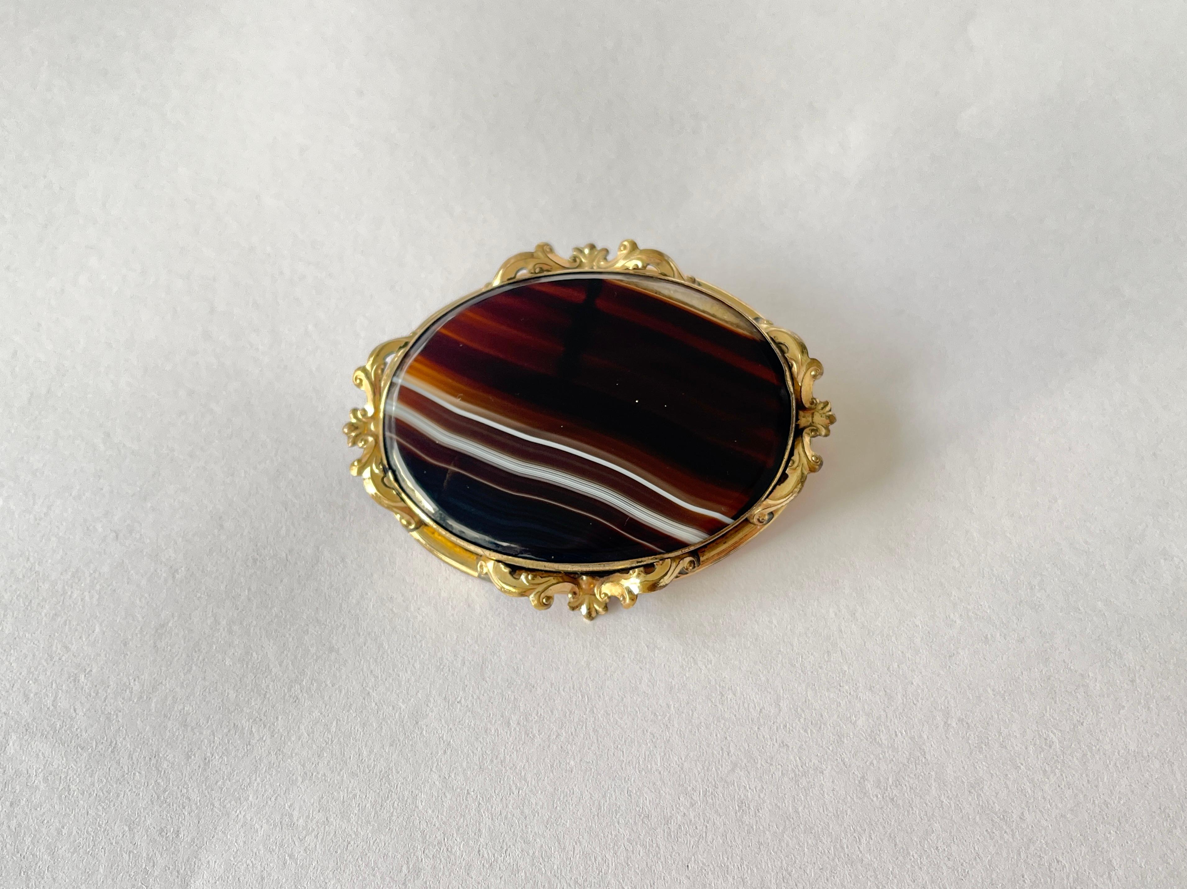 This lovely brooch is big and bold!
It features a piece of brown, caramel and white striped banded agate that is oval in shape.  It is set in gold gilt with nice detail of the fleur de lis and scrolling leaves.  
This is a large piece measuring over
